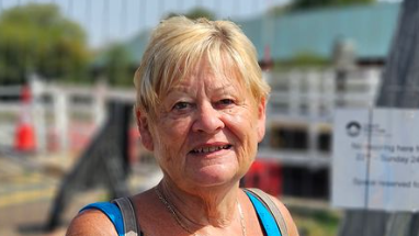 A woman in her 70s with short blonde hair smiles as she stands in front of a swing bridge which has been closed off with barriers for repair work. The blue straps of her vest top and the grey straps of her backpack are visible.