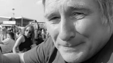 A black and white selfie showing the face of a man with blond hair. Blurred figures are visible behind him.