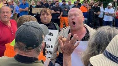 A bald man holding a placard is shouting at a man who has his back to the camera. There are lots of other people around, some holding cardboard signs, across the road