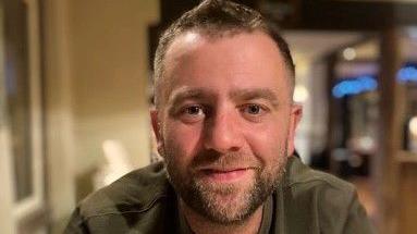 Scott Bryan, a man with short brown hair and a stubbly beard, is wearing a dark green t-shirt.