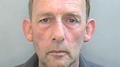 Mugshot of Brian Furneaux who was sentenced to 13 years in prison. Furneaux is looking directly at the camera. He is a white man with light brown hair and dark eyes. He is wearing a dark-coloured top and is standing in front of a light grey wall. 