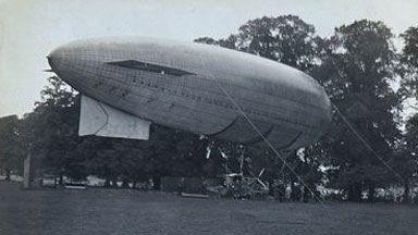 A black and white image of a Beta II airship floating above grass at Jesus Grove Cambridge, in 1912. It is tied to the ground with ropes. Behind it is an avenue of trees.