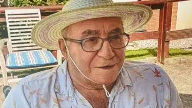 An elderly man wearing glasses and a sunhat, while wearing a white shirt. He is sat in a chair outside looking off to the right of the camera