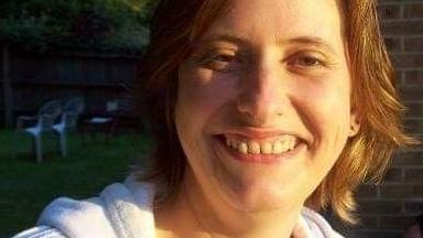 A woman dressed in a white hoodie smiles at the camera. He face is lit up by the sunshine and she has a brown shoulder-length hair.