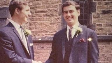 A wedding photo taken in the 1970s of two men in navy suits with white flowers on their lapels.