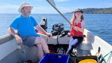 A small girl sits in a small boat with a middle aged man. The boat has an outboard motor and has an island behind. The girl holds a flag.