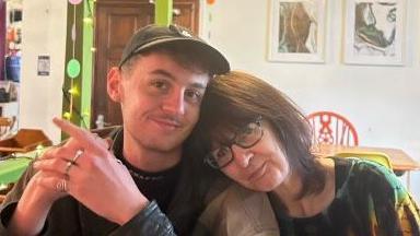 Cai Morgan sitting beside his mum, in a cafe, who has her head on his shoulder. He is wearing a dark top and jacket with silver rings. He is smiling at the camera. She is wearing a green and yellow top, with dark glasses.