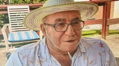 An elderly man wearing glasses and a sun hat, while wearing a white shirt. He is sat in a chair outside looking off to the right of the camera