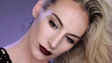 Një grua me flokë bjonde pozon për kamerën. Ajo e ka kokën të anuar anash dhe flokët e saj janë të hedhura mbi kokë dhe varur poshtë saj. Ajo ka grim të errët, duke përfshirë buzë të kuqe të errëta dhe laps sysh të zi.