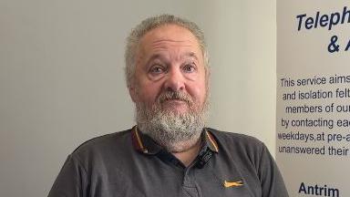 A man with grey hair and a grey beard looks towards the camera. He is wering a dark grey polo shirt with yellow details.
