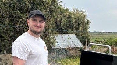 Jake Carroll smiling for a photo while making an outdoor barbecue in the countryside. He is wearing a dark baseball cap and white vest top. A garden shed and trees can be seen in the background.