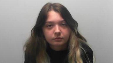 A young woman with long dark hair featuring blonde streaks sits against a plain light‑coloured wall, wearing a black sweatshirt.
