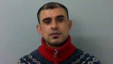Salam Karis has short dark hair and is wearing a red knitted jumper. The image is a mugshot, so the wall behind him is grey.