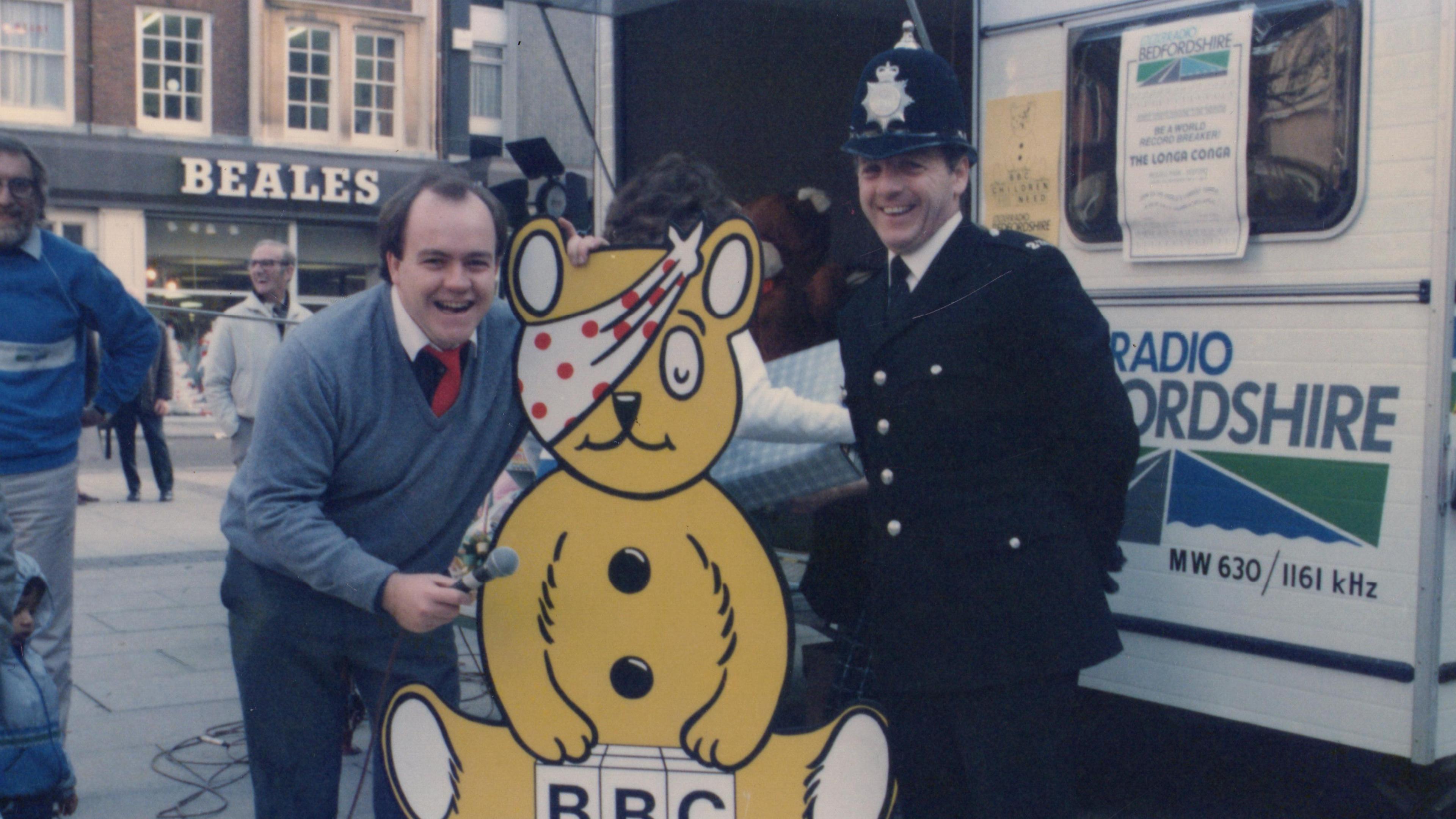 Gerald Main holding a microphone while standing next to a cardboard cutout of the yellow BBC Children in Need teddy bear and a uniformed police officer. They are on the street in a town centre.