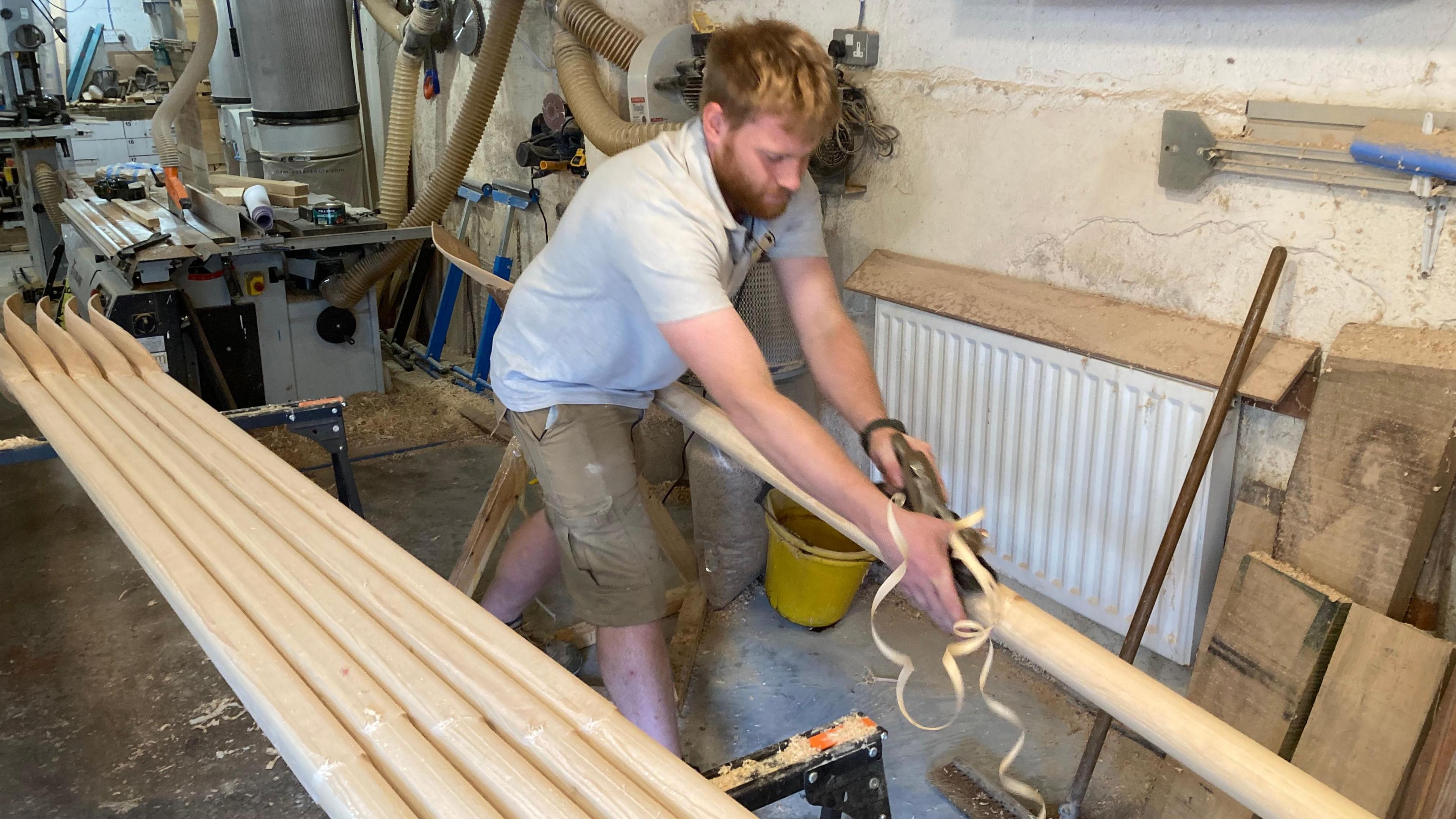 A bearded man with ginger hair in a grey polo shirt working on a wooden oar in his workshop with a slice of wood falling to the floor.