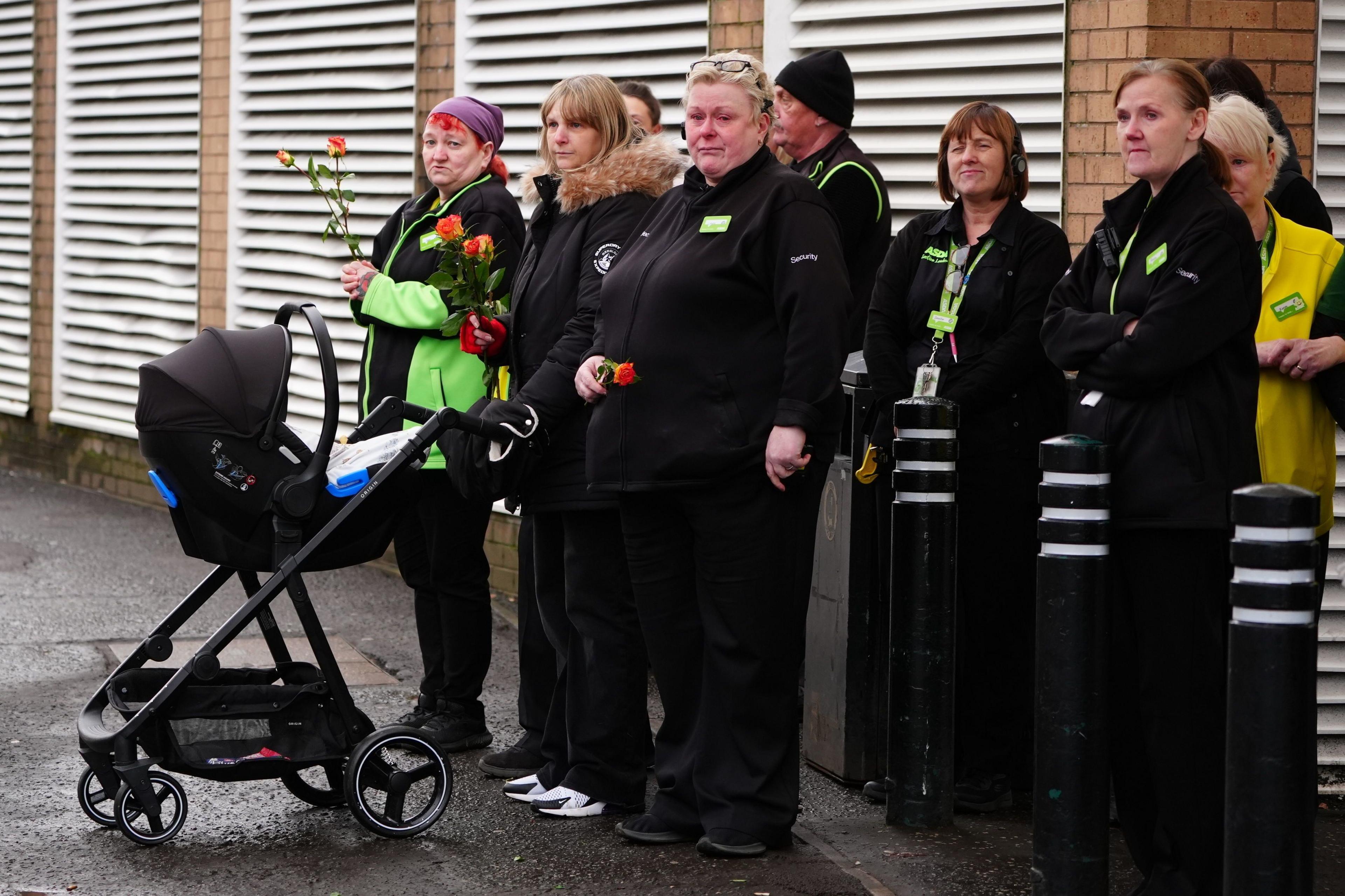 A number of workers in Asda uniforms stand on a pavement. Some are tearful, others are holding orange roses. One has a pram.