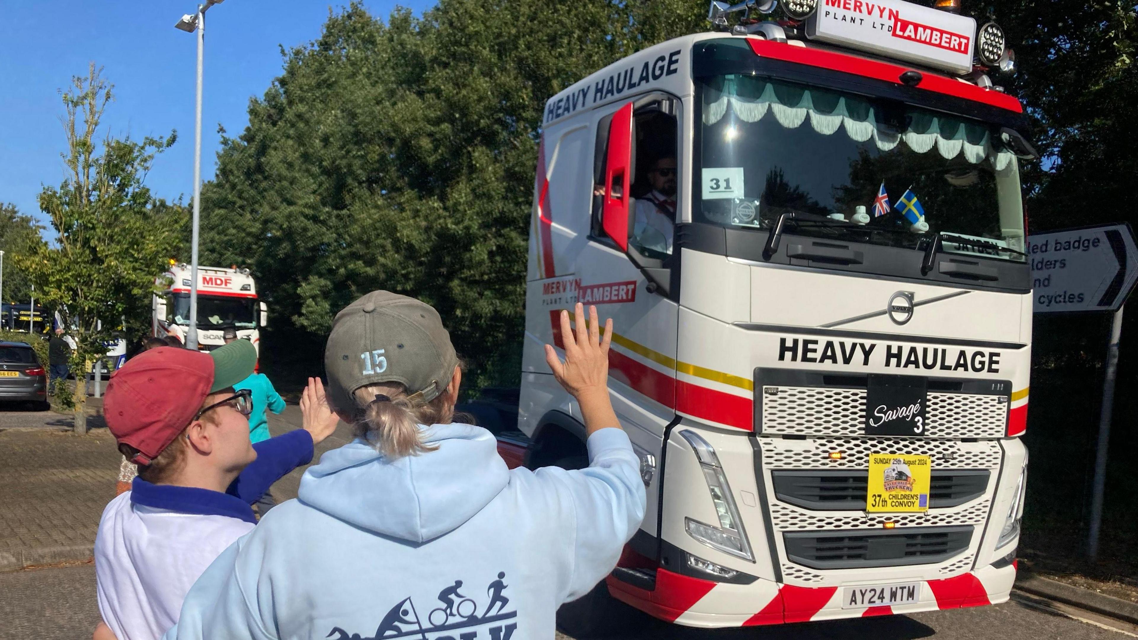 Lucy and Samuel, who are arm in arm, wave at a truck