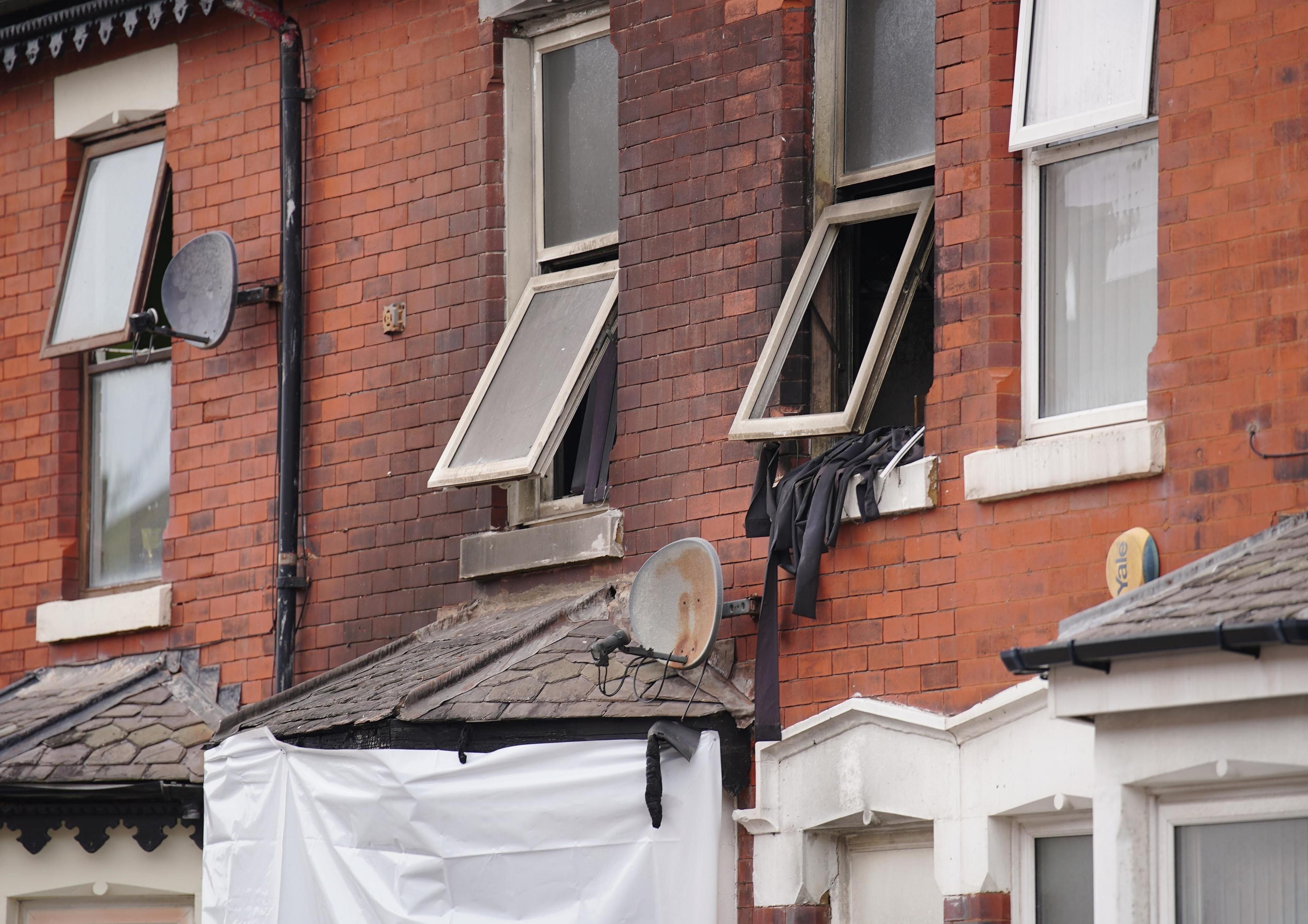 A white sheet covers the window of the burned out house after a fatal blaze. Two open windows are open and the upper half of the house is blackened and some blinds appear to be hanging out. 