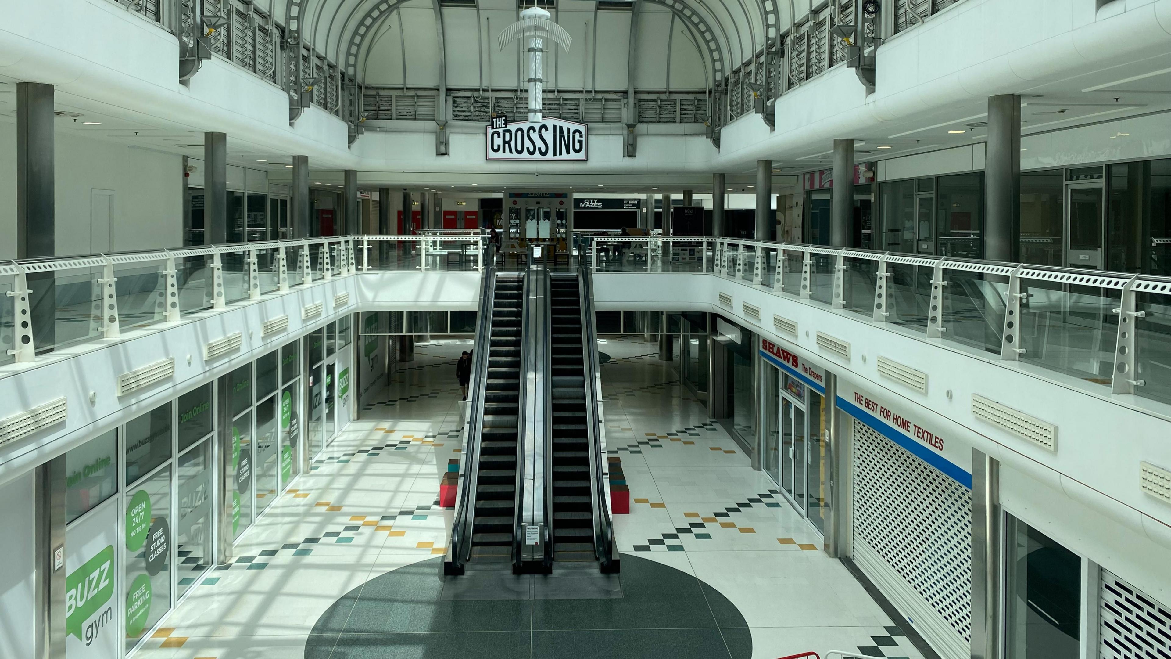 The inside of the Brunel shopping centre in Swindon taken from the first floor. There are a number of empty shop units. In front there are empty escalators. There is a sign saying "The Crossing". There are no people in the shot.