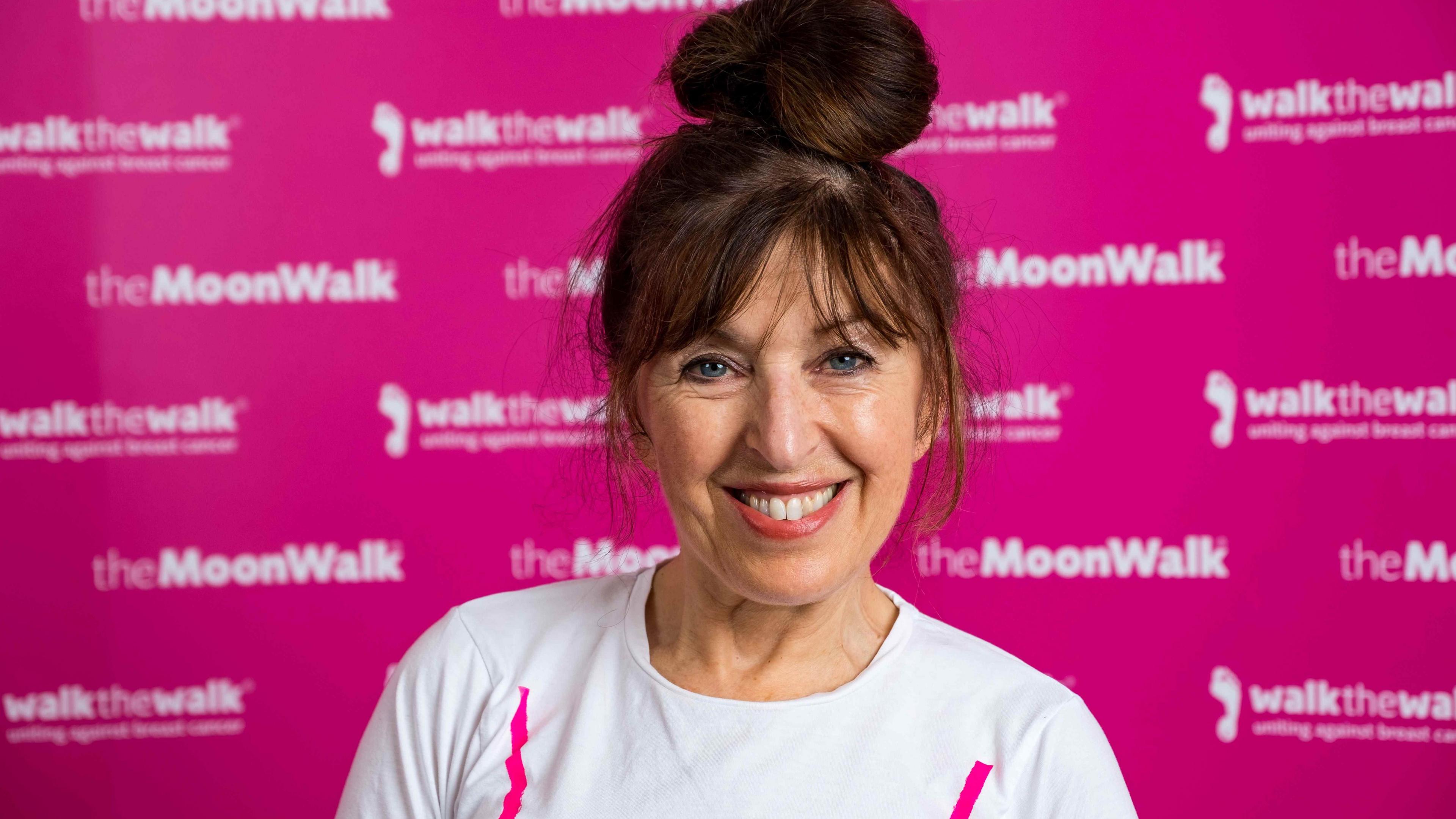 Nina Barough smiling at the camera, has dark hair, blue eyes and is wearing a white t-shirt. Behind her is a pink board saying the Moon Walk.