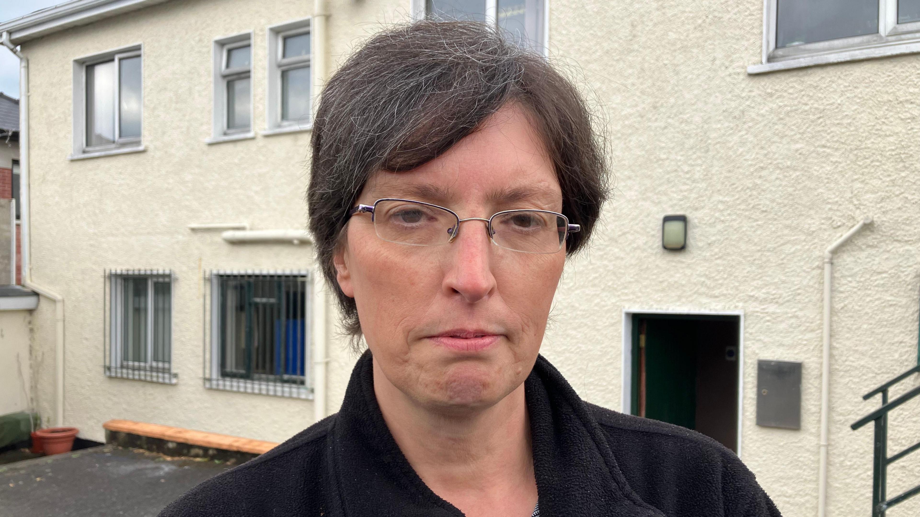 A lady with short dark hair stands in front of a white building. Sh has a sombre expression, is wearing eye galssses, a black coat and grey and white top