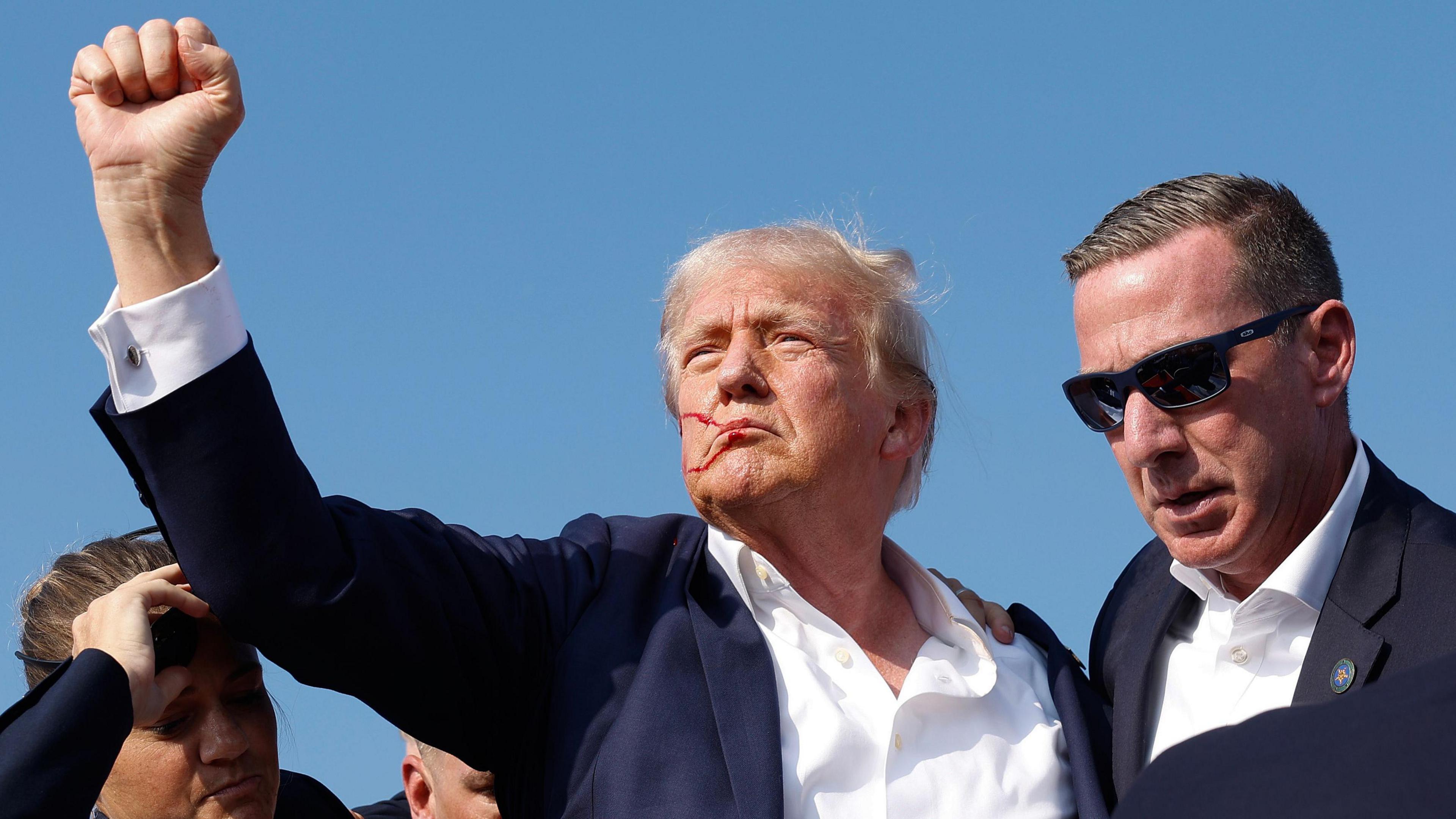 Donald Trump holds up his fist as blood is smeared across the left hand side of his face. He is flanked by two secret service agents.