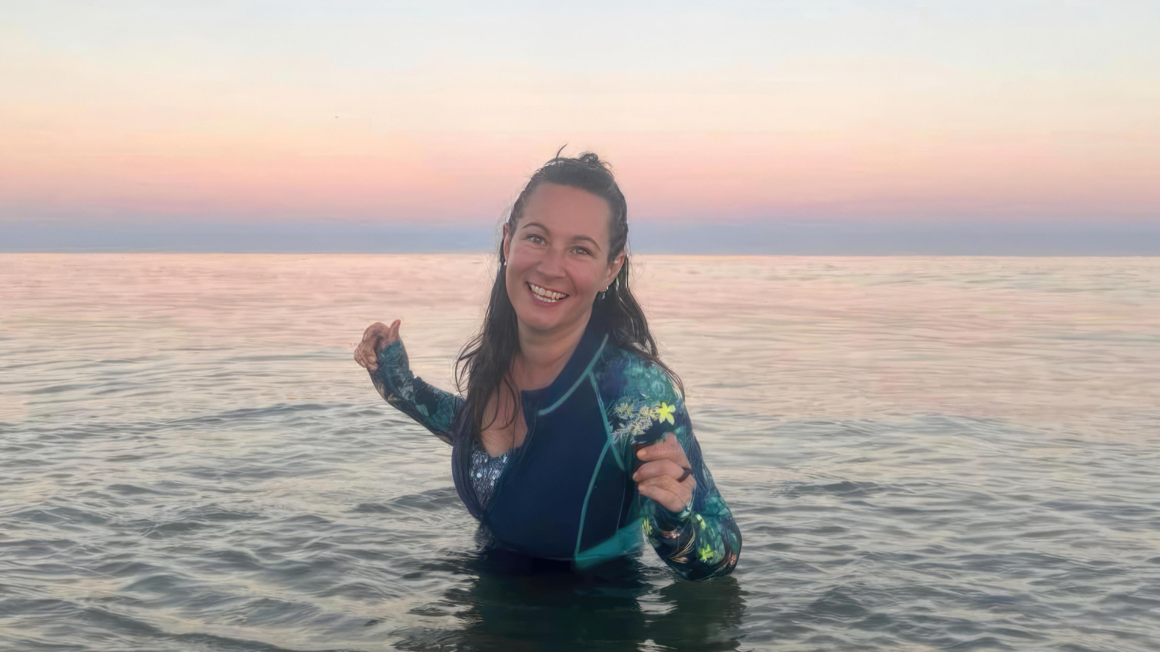 A woman in a wet suit standing on the sea, the sun setting behind her