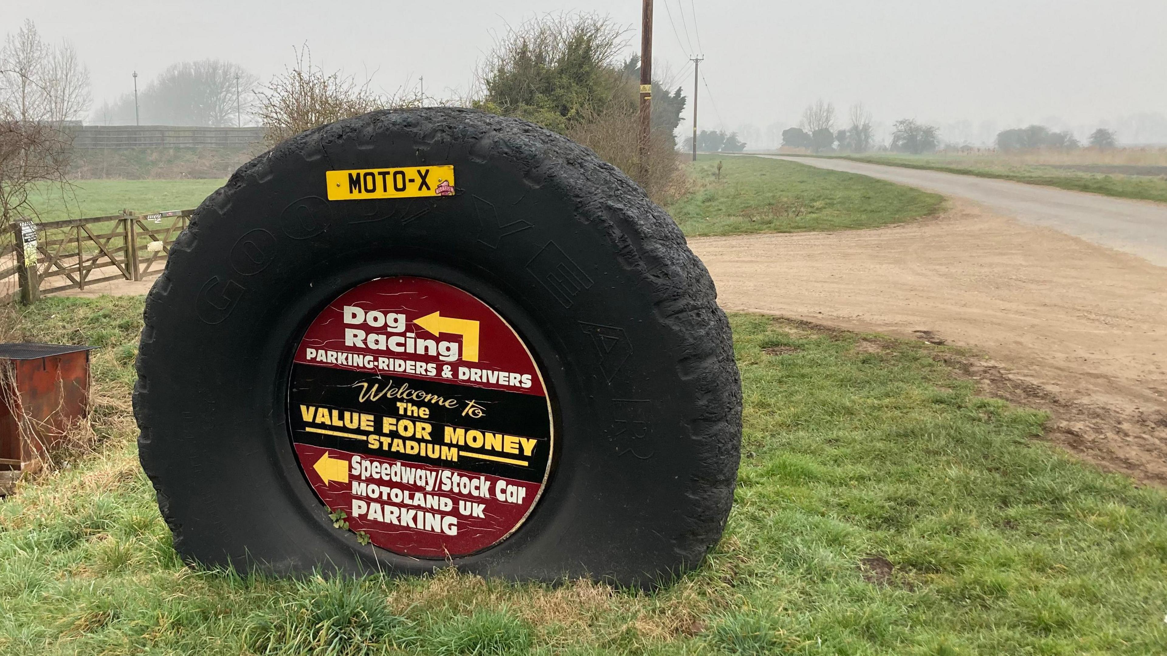 Large tyre embedded in grass. Its centre advertises the motocross track and events. 