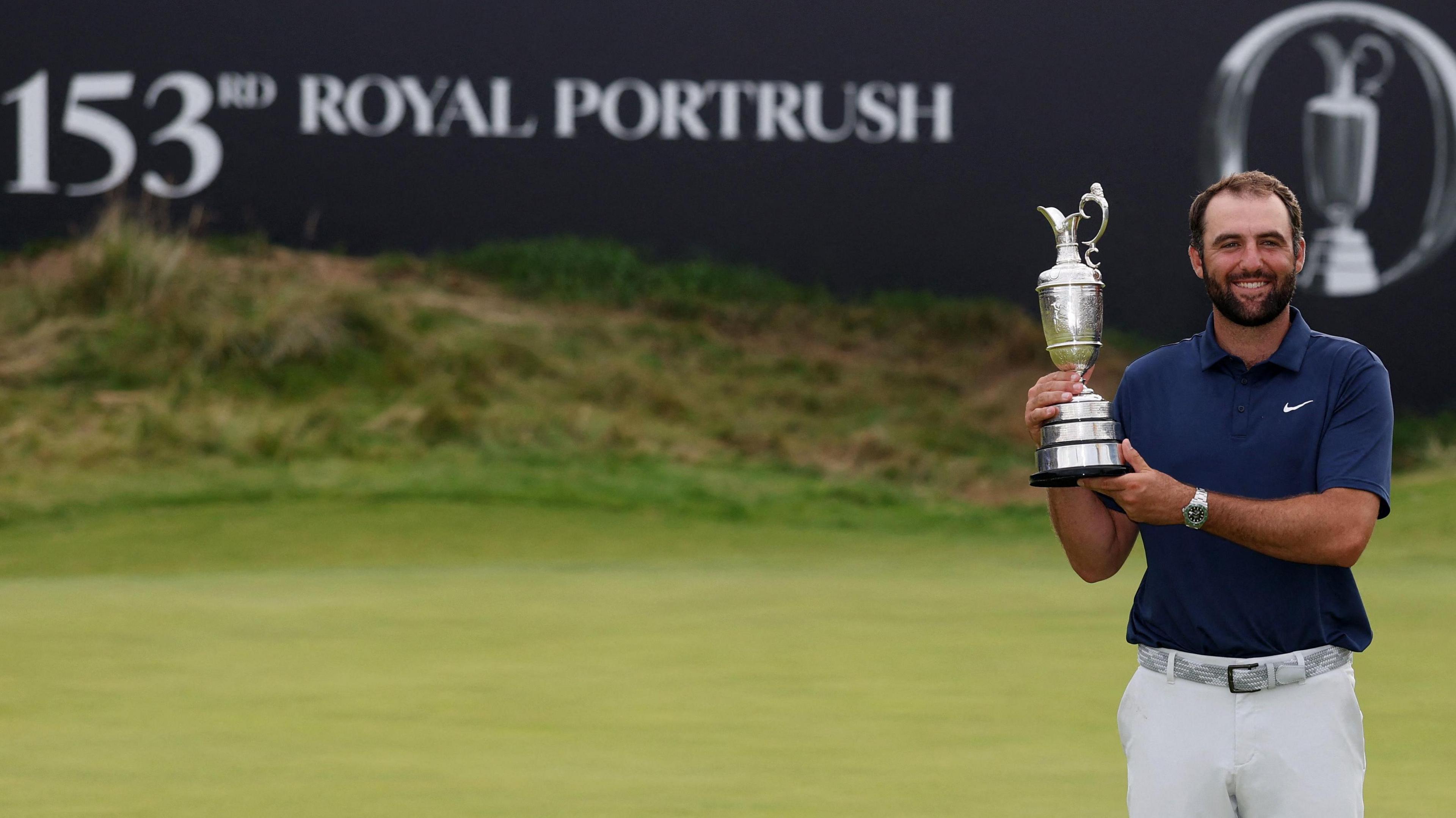 A man in a navy blue polo shirt, white trousers and a silver wrist watch holds up a trophy up and smiles at the camera. Behind him is a putting green and a sign which says "153rd Royal Portrush".
