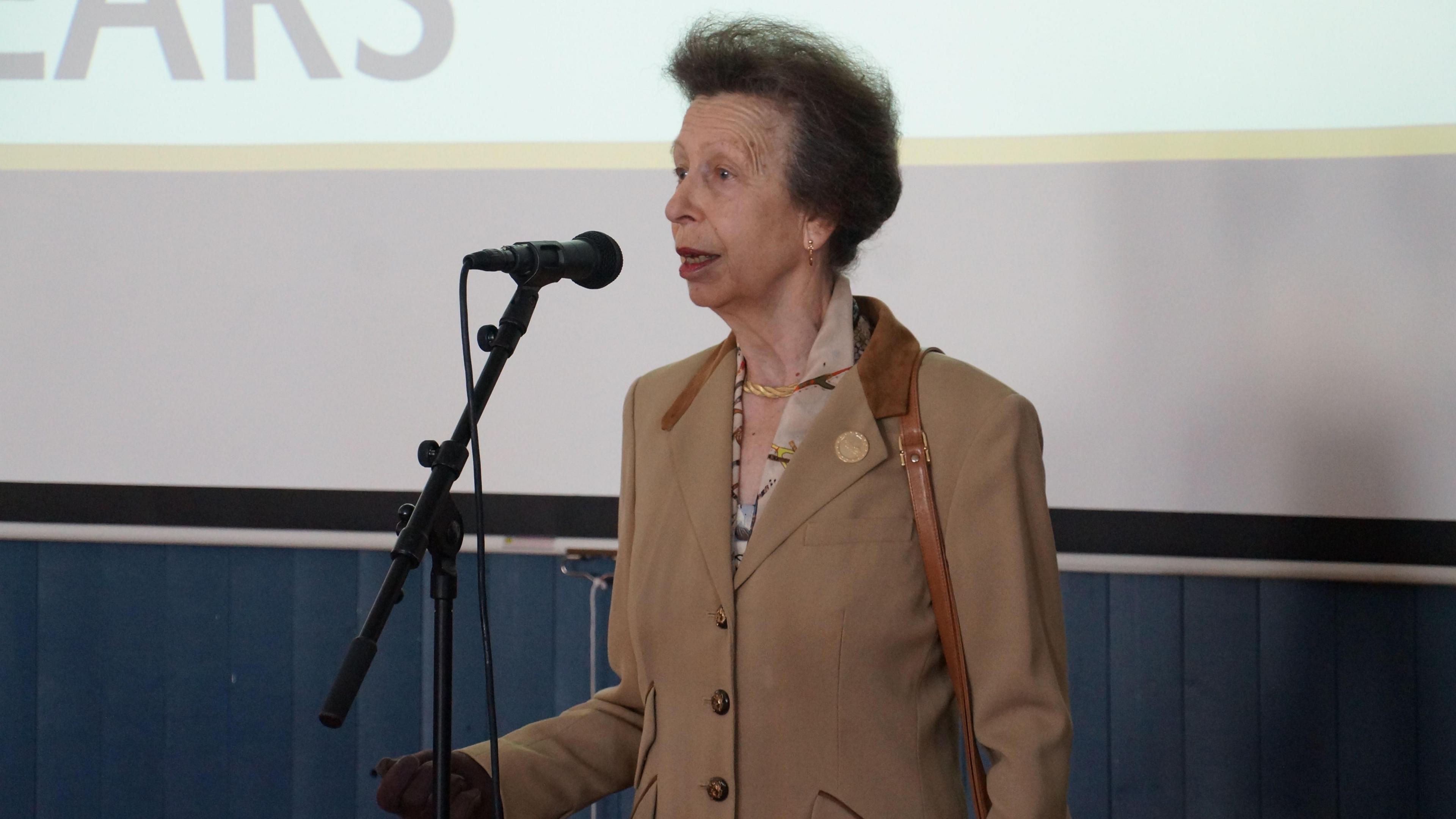 Princess Anne speaking into a microphone fixed on a stand, with the lower section of a large projector screen behind her. She is wearing a camel-coloured blazer and brown gloves.