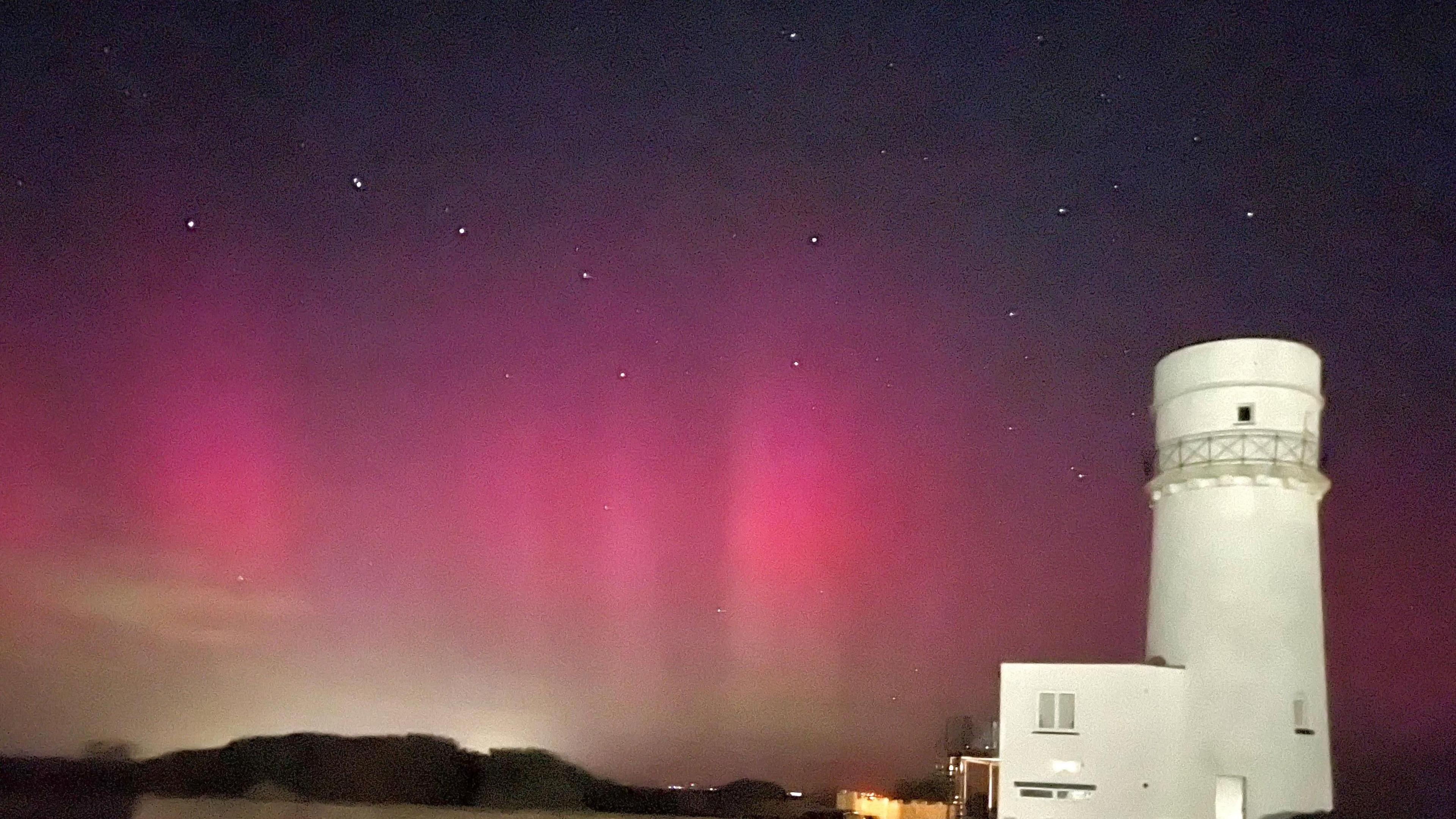 A white lighthouse is pictured to the right against a night sky that is brightly lit with pink light.