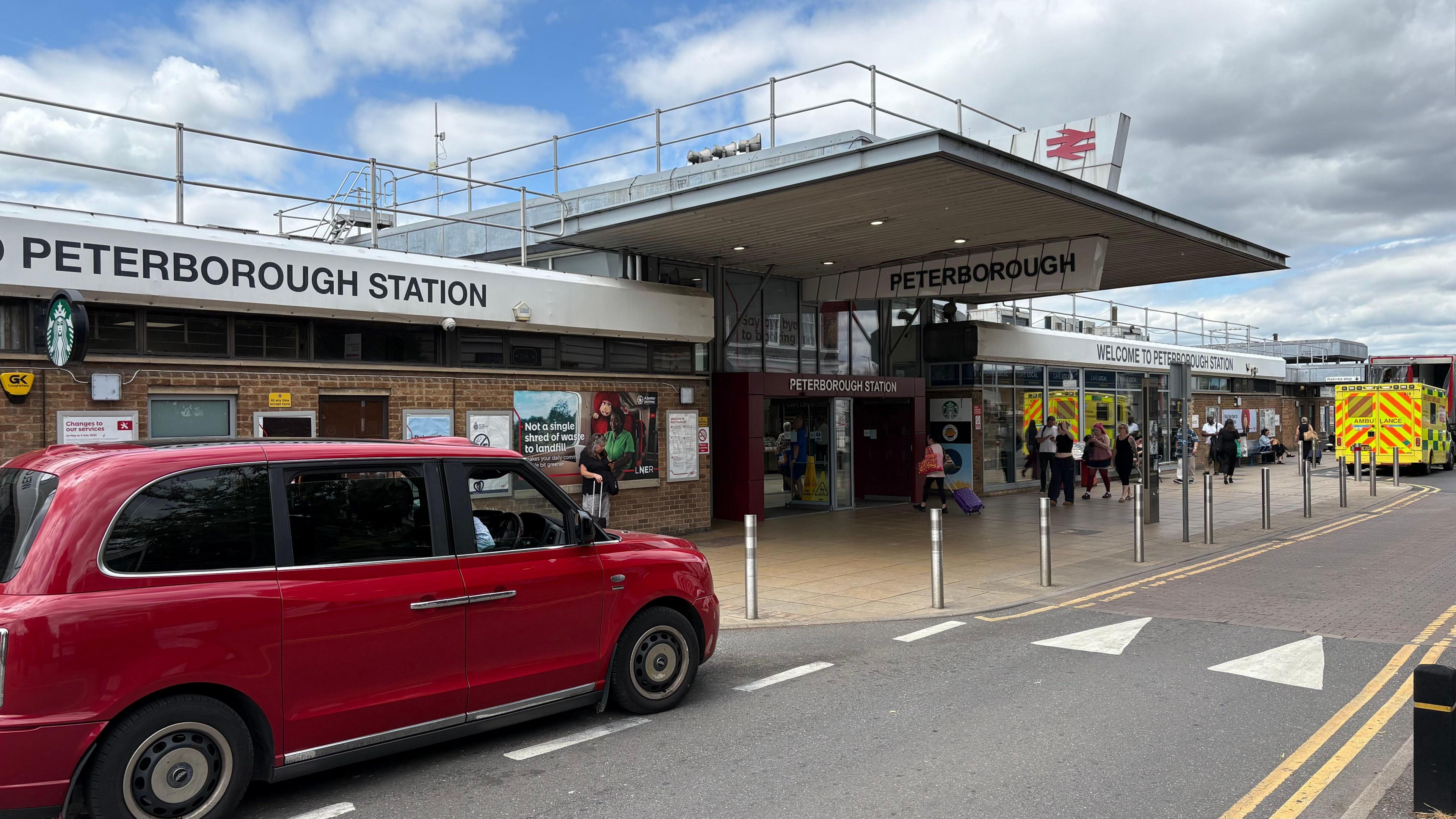 Outside of Peterborough Station on a day with blue sky and some clouds. People are walking into the small entrance way. There are taxis parked at the front as well as an ambulance.