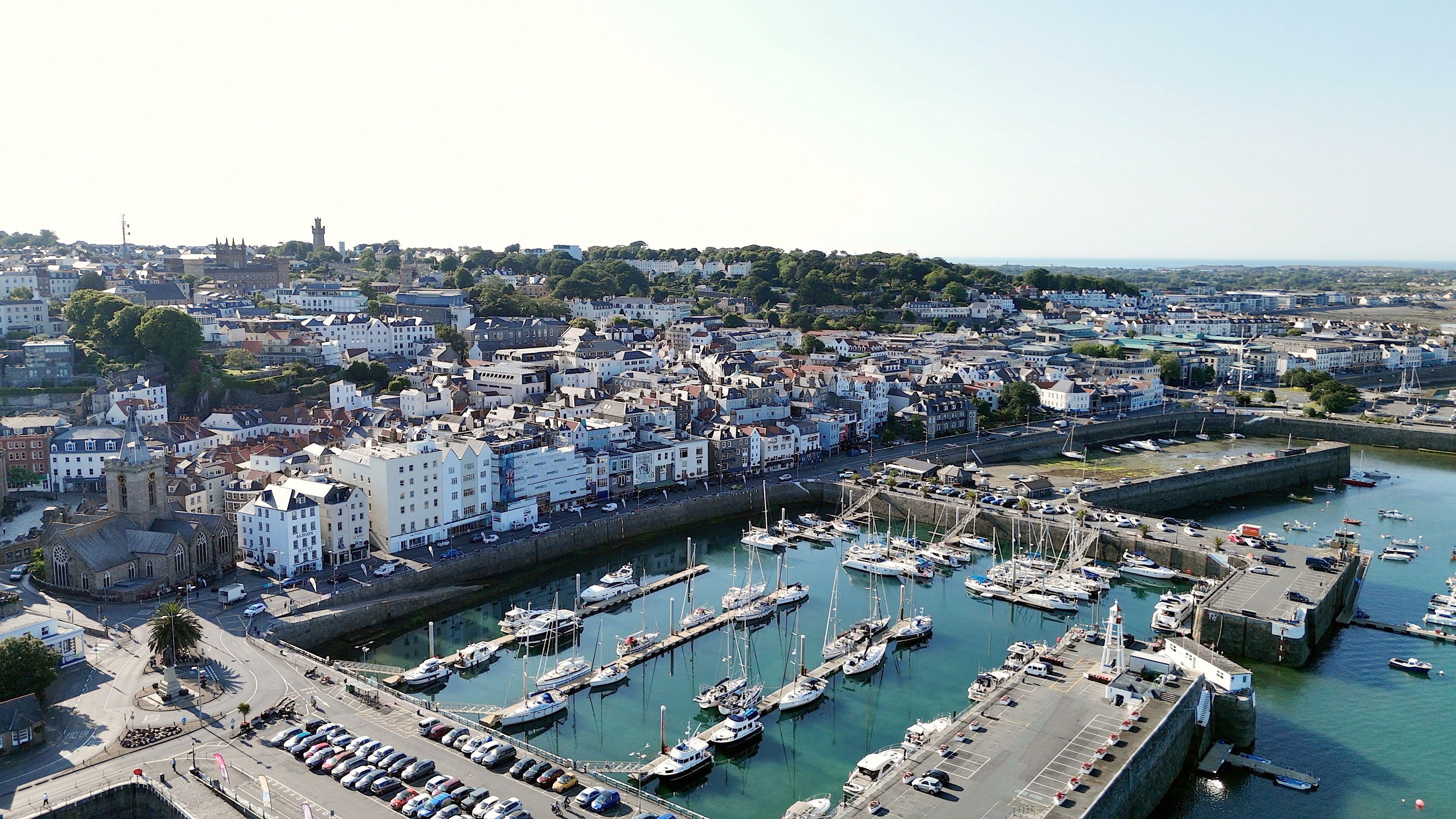 An aerial view of Guernsey's main marina in St Peter Port. There are dozens of boats and yachts moored to piers in the marina. The town rises gently from the waterfront. 