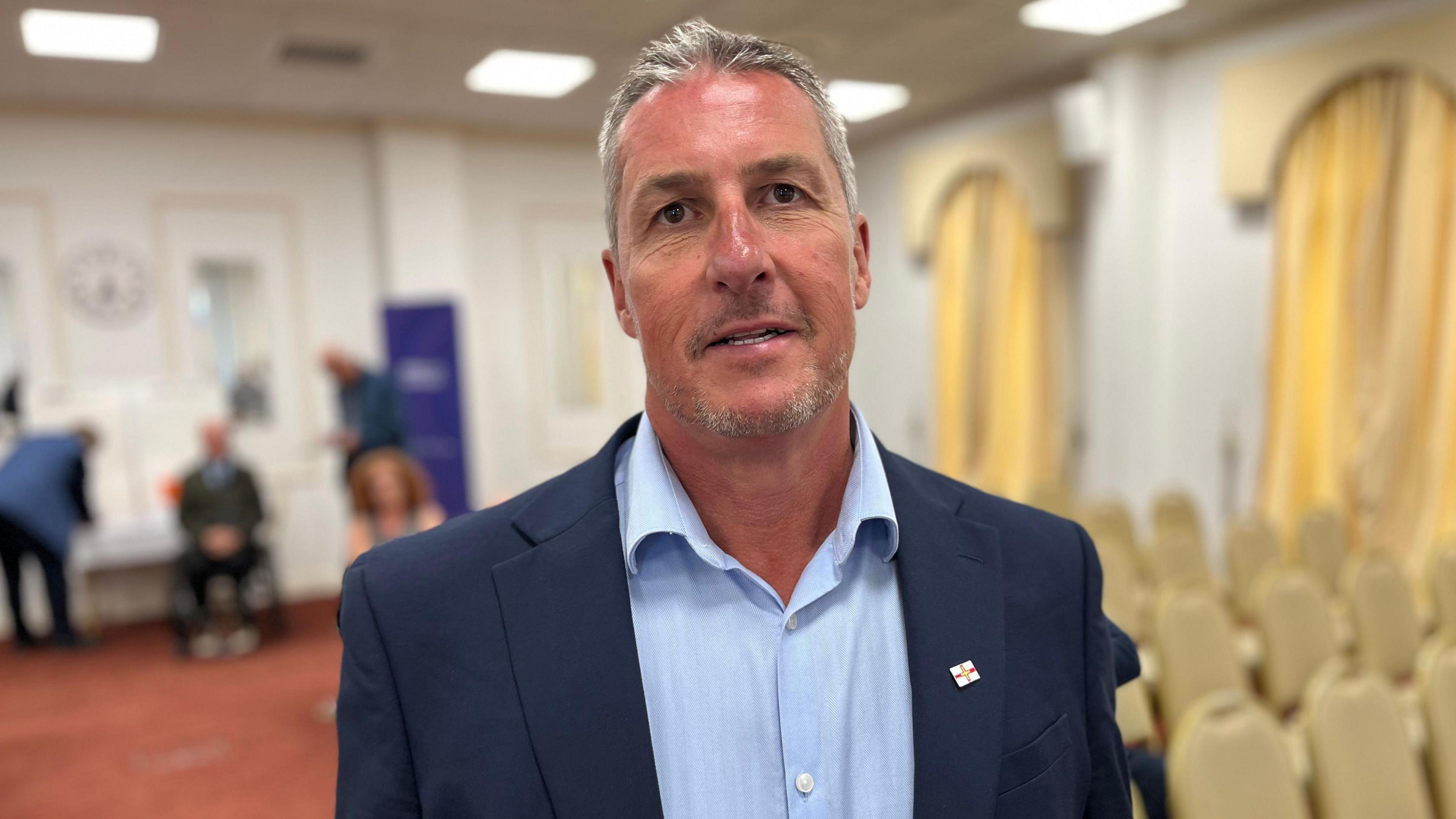 Deputy Andy Cameron - A man with grey hair, wearing a blue suit and a blue shirt with a Guernsey flag on his lapel. 