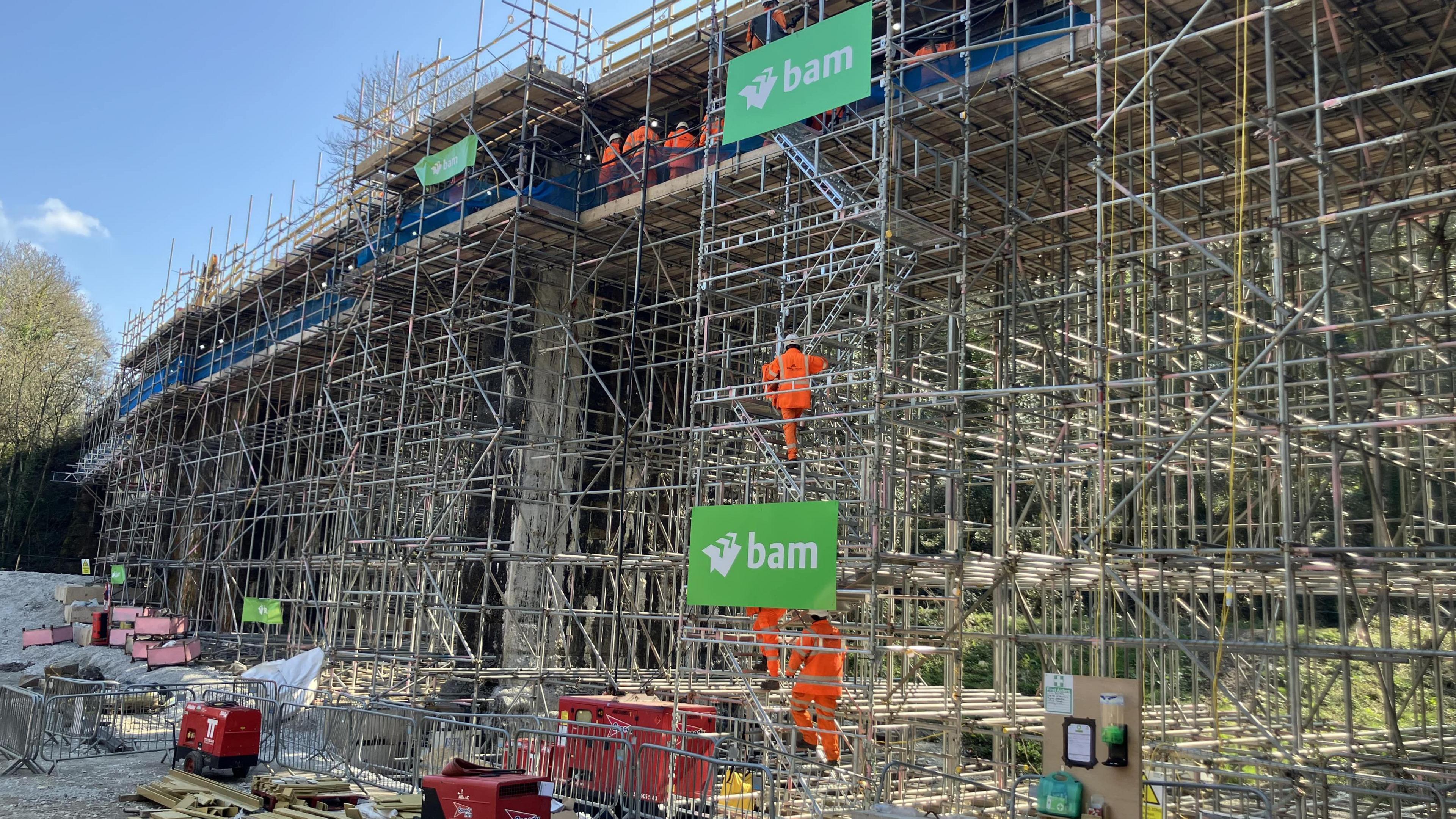 The image shows a lot of building and construction work going on, with lots of scaffolding. There are three workers dressed in orange hi-vis clothing climbing the scaffolding. There are also generators and fencing on the ground. 