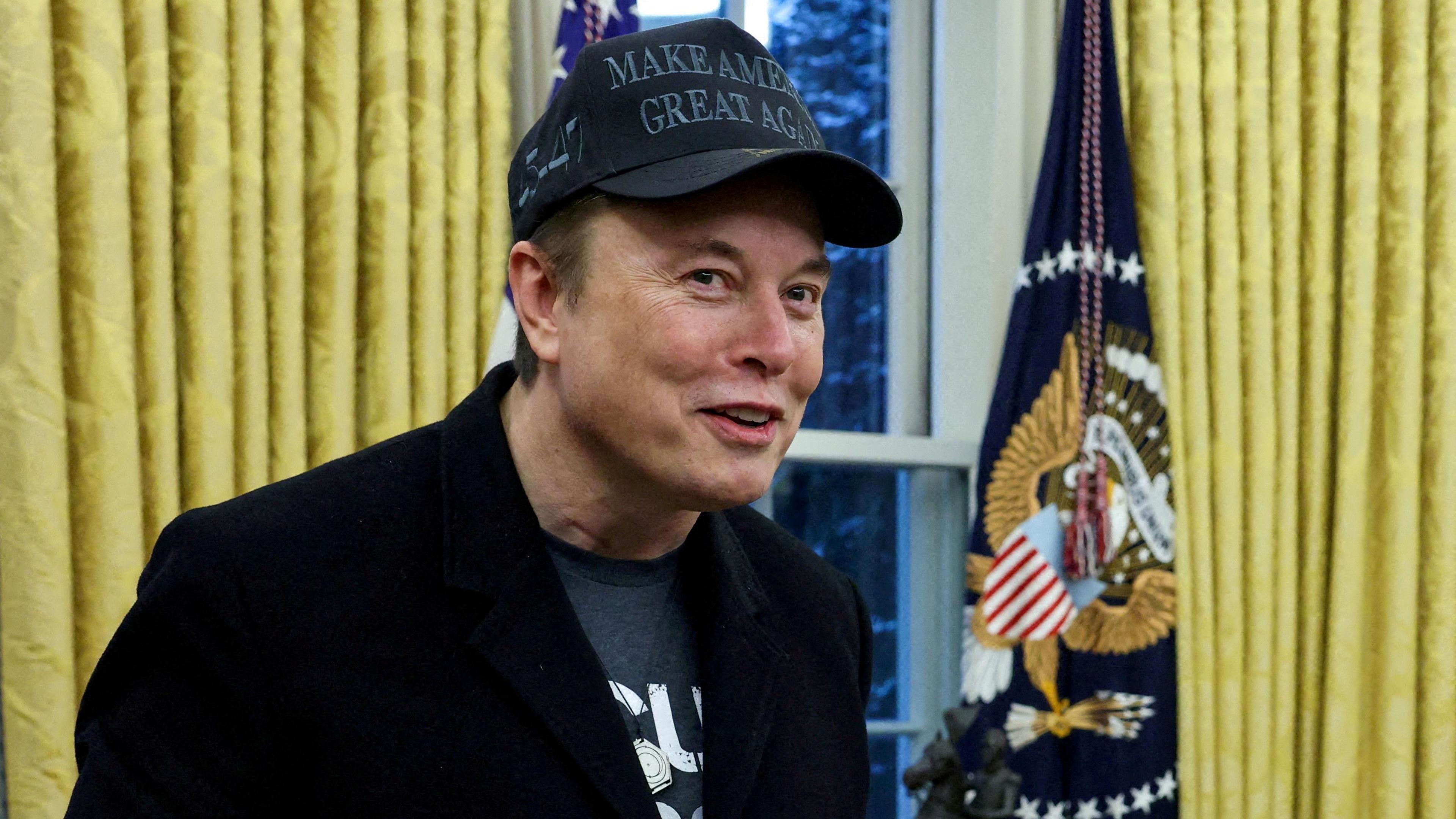 Elon Musk, a man with dark hair covered by a black "Make America Great Again" cap, smiles in the Oval Office. He is wearing all black and visible from the chest up. Behind him are gold curtains, a window and a US flag. 