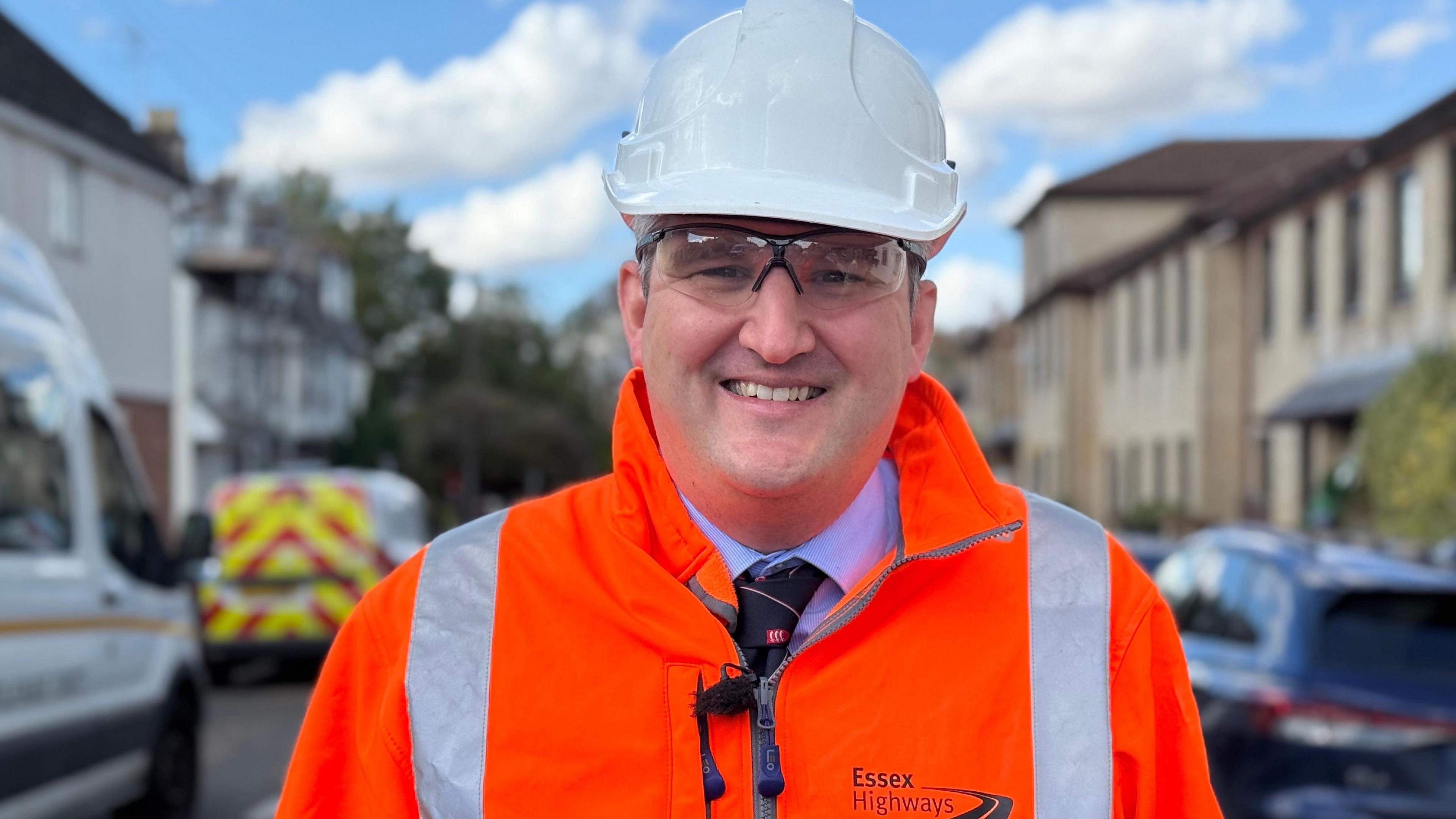 Councillor Tom Cunningham is smiling at the camera. He is wearing an orange hi-vis jacket, goggles and a white hard hat. He is standing on a residential road where road repairs are taking place. Highways vehicles can be seen on the left.