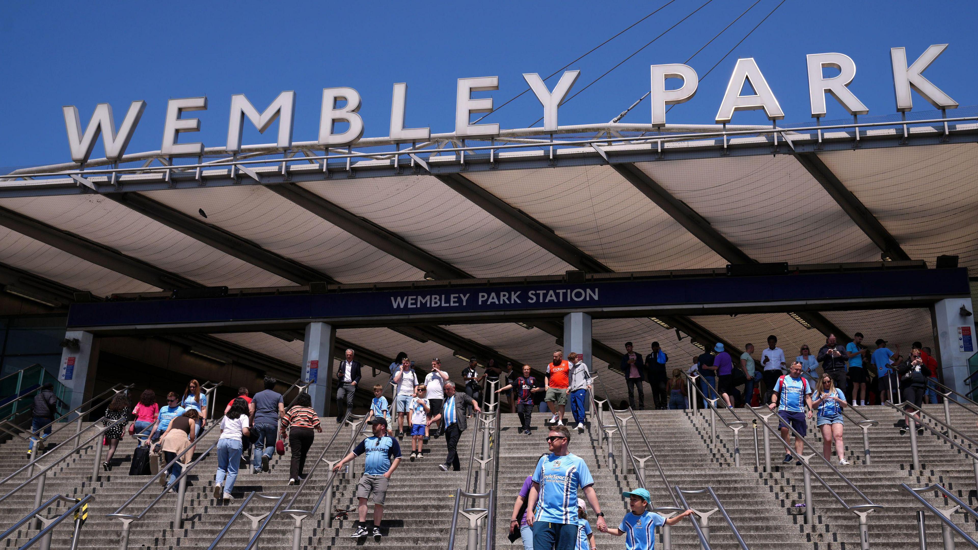 Football fans walk down stairs from Wembley Park station towards Wembley Stadium. It is clearly sunny day. The fans are most wearing sky blue Coventry City football shirts.