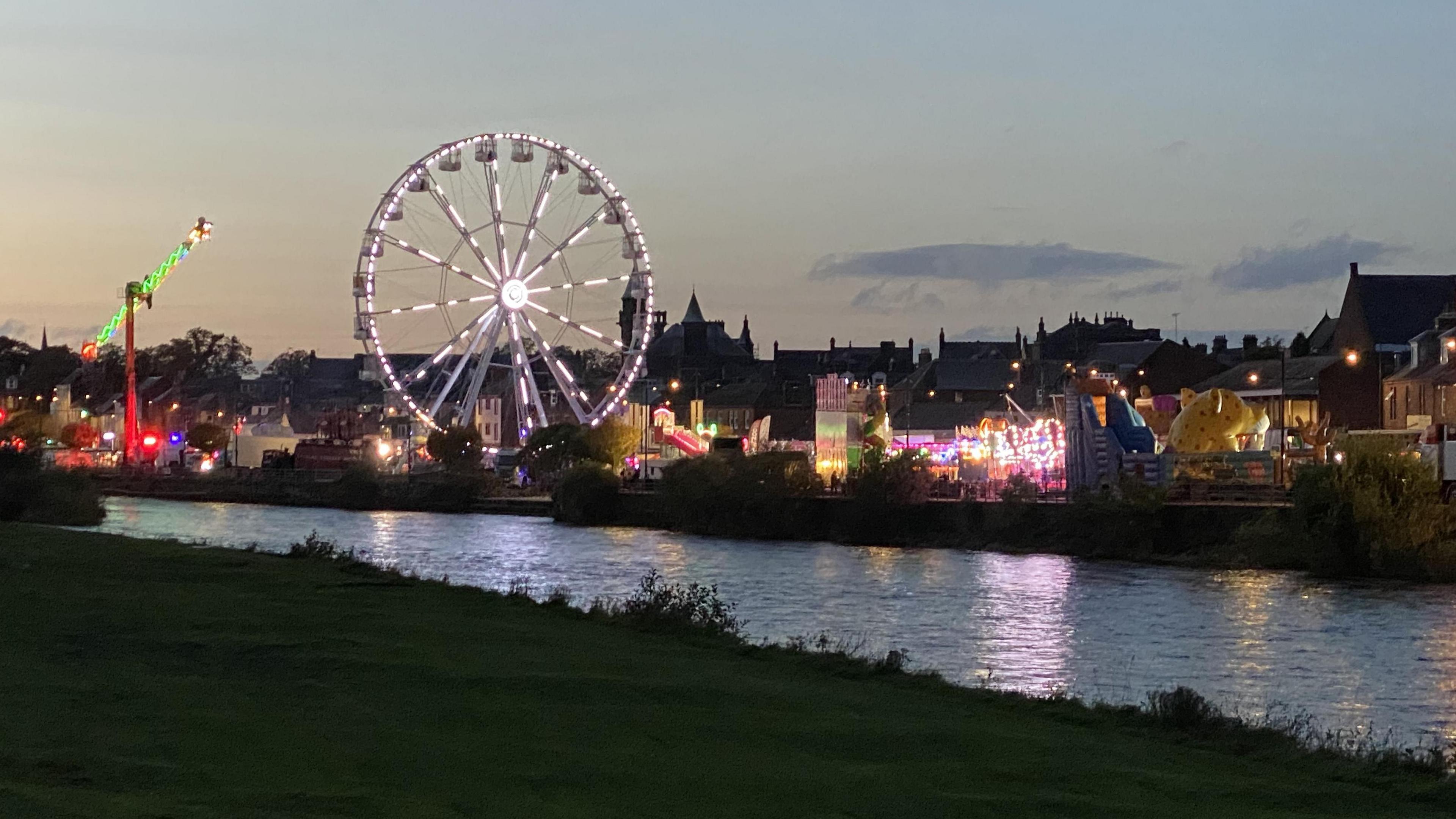 A fair viewed across the River Nith in Dumfries at night with a big wheel and other riders lit up over the water