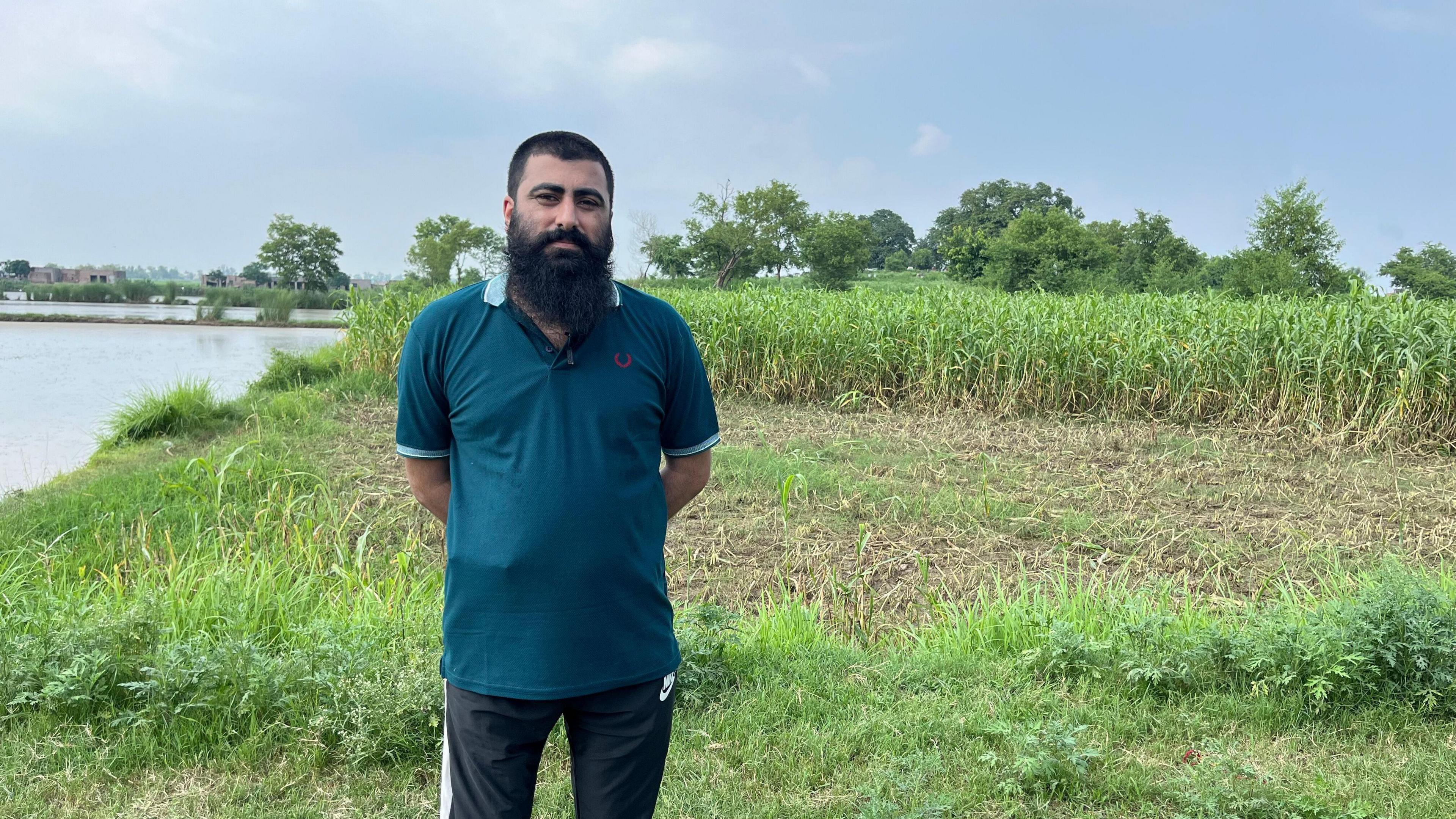 Mr Munir dressed in a blue T-shirt and black tracksuit pants stands next to a field of corn which is next to a body of water.