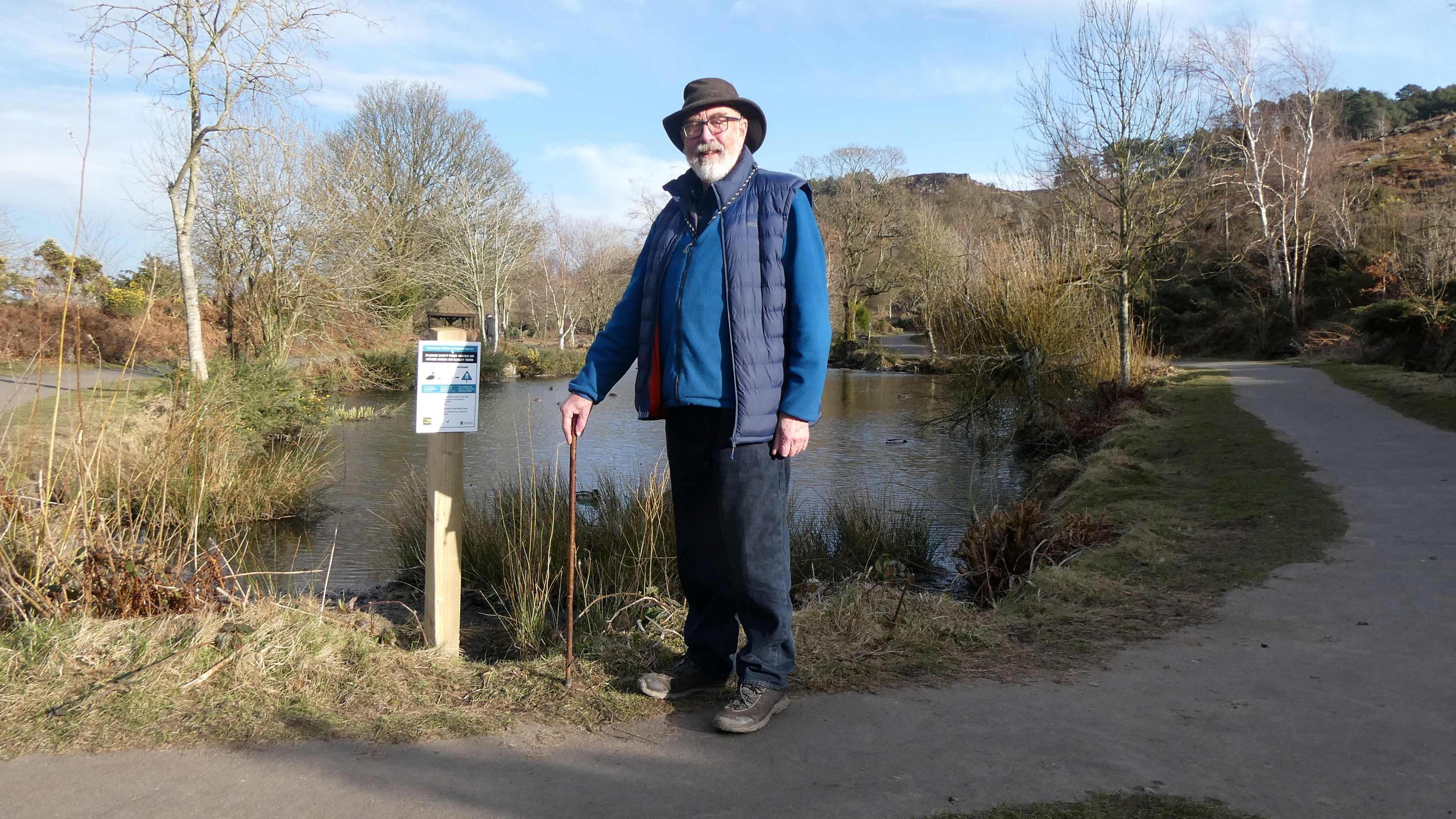 Owen Wells, chairman of Friends of Ilkley Moor, stands next to a notice by a lake discouraging feeding the ducks and other birds. A walking stick is in his right hand, he has a white beard and is wearing a blue jumper and gilet. 