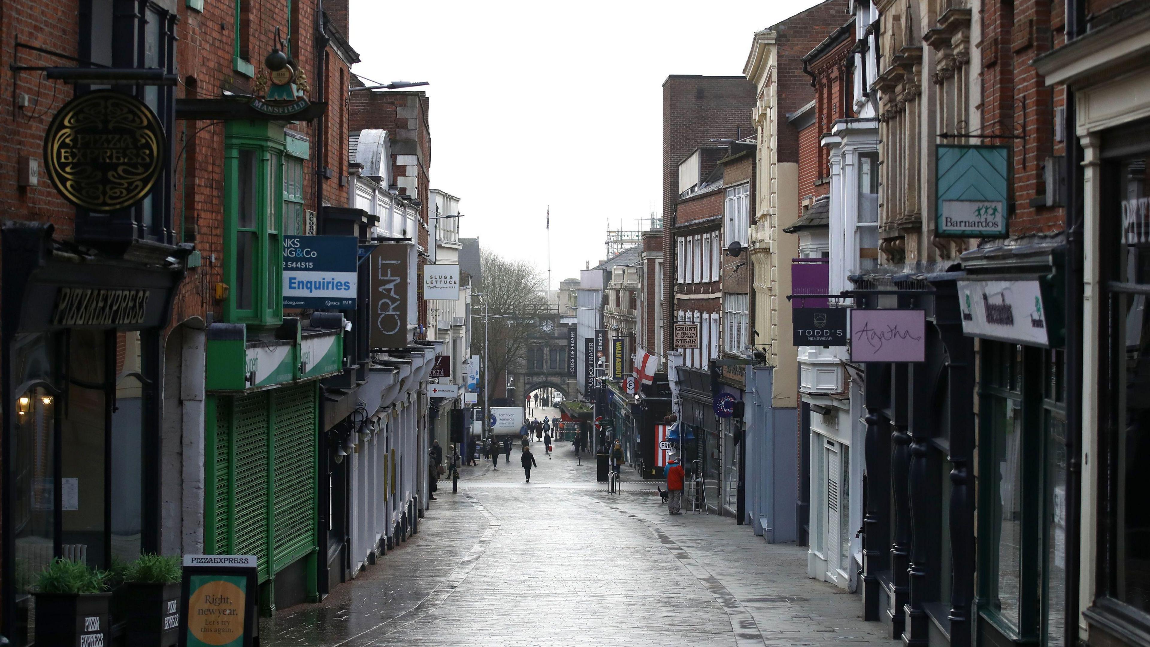 A view of a shopping street looking downhill. There is a row of shops on either side of the cobbled street. The shops are closed with shutters down on a few of them.