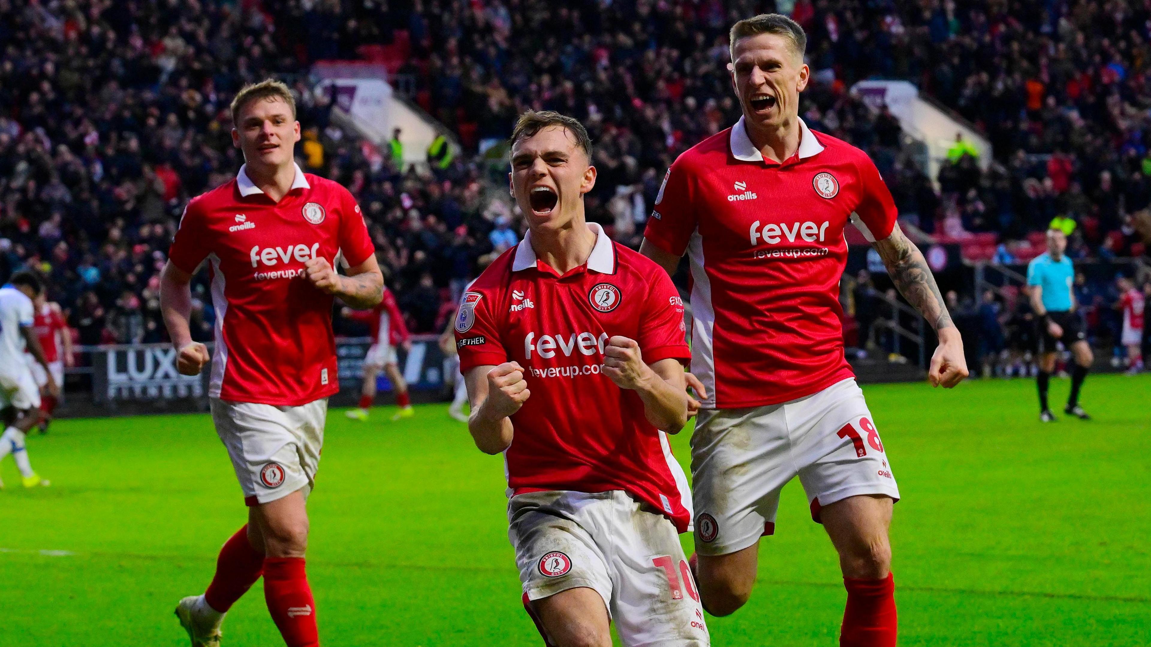Scott Twine (centre) clenches both his fists and has his mouth open as he shouts in celebration after scoring, with Emil Riis (right) and Cameron Pring (left) behind him