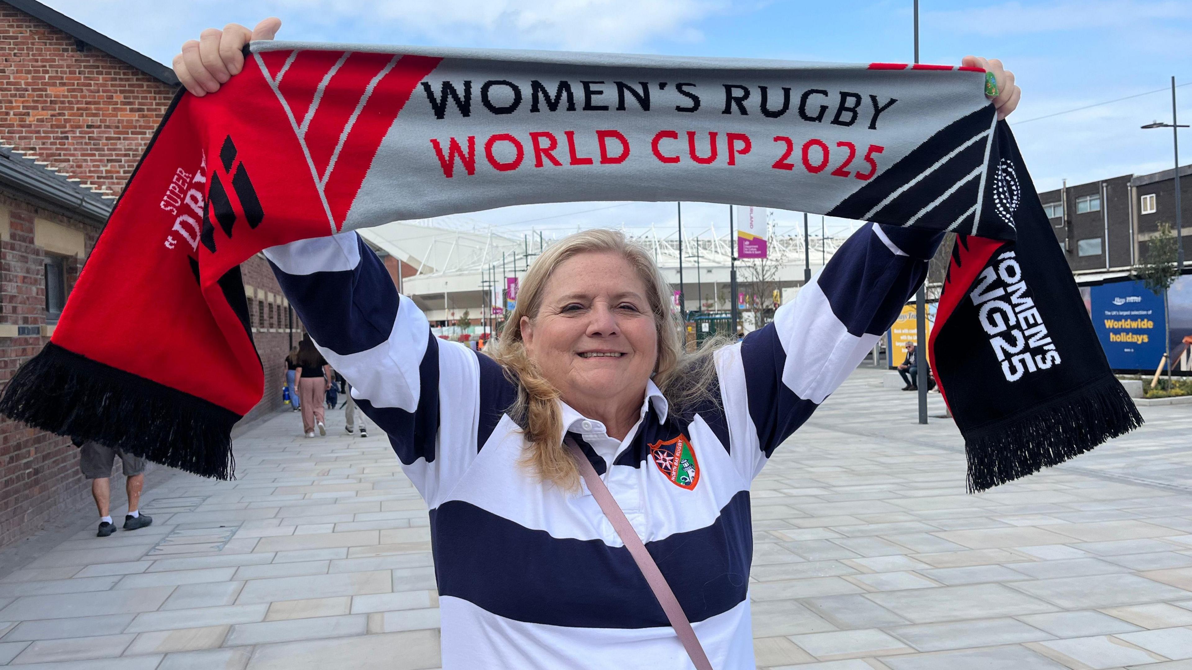 A woman wearing a striped white and blue rugby shirt holding her arms aloft and smiling. She is holding a banner proclaiming "women's rugby world cup 2025". Behind her the roof of a stadium can be seen.