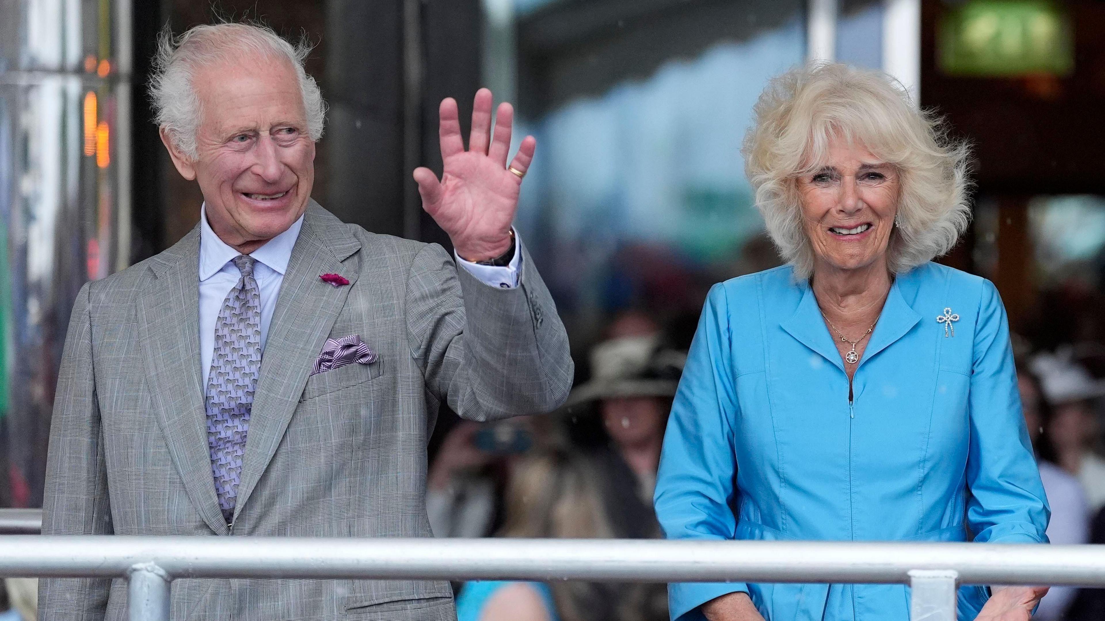 King Charles III and Queen Camilla stand on a platform while on a royal visit to Jersey. The King has a grey suit on and is waving to crowds with his left hand. The Queen has a sky blue dress on.