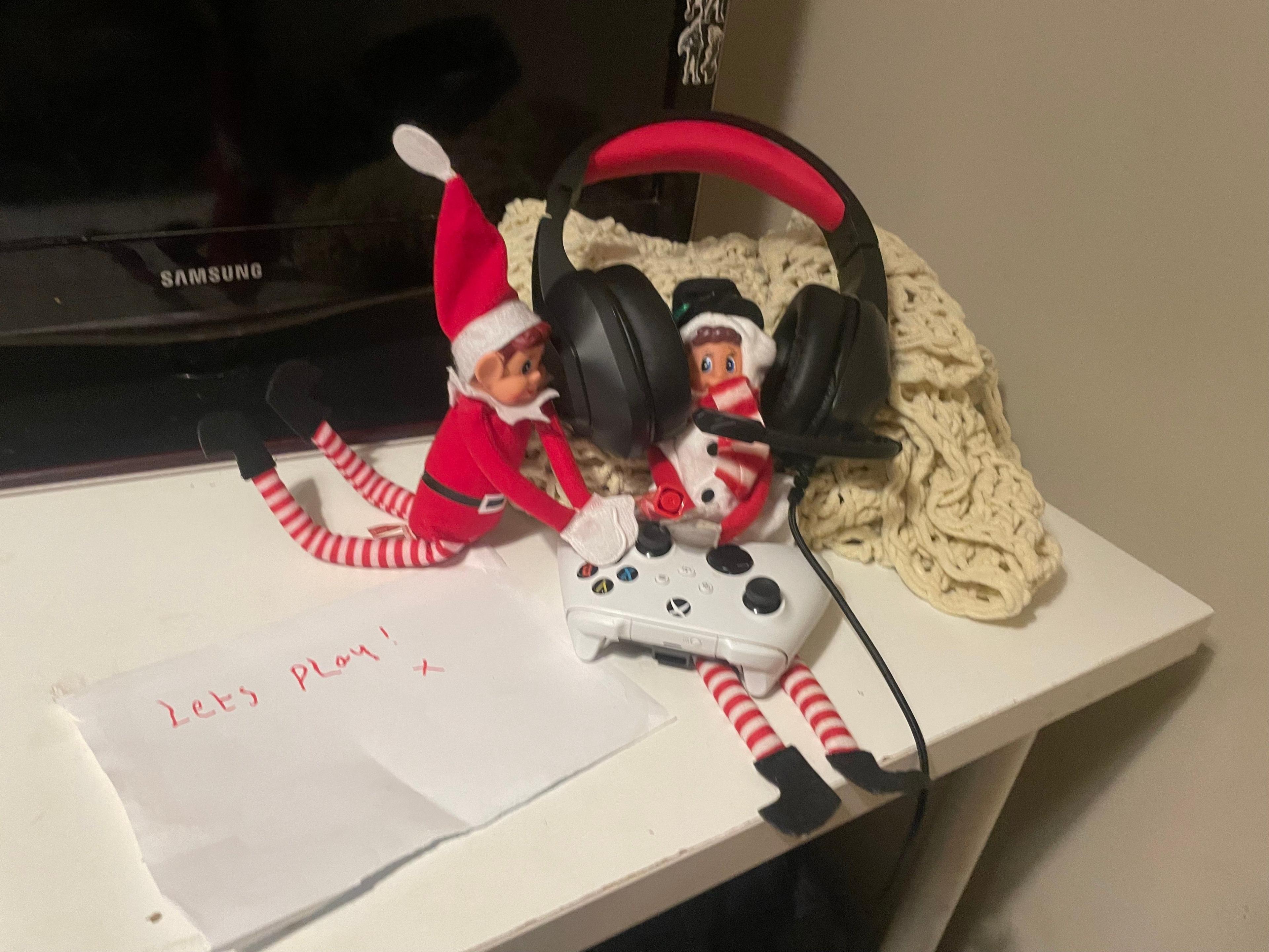 Two elves sat on a table. One has headphones on and is sat with a controller on its lap. Another elf is trying to press buttons.