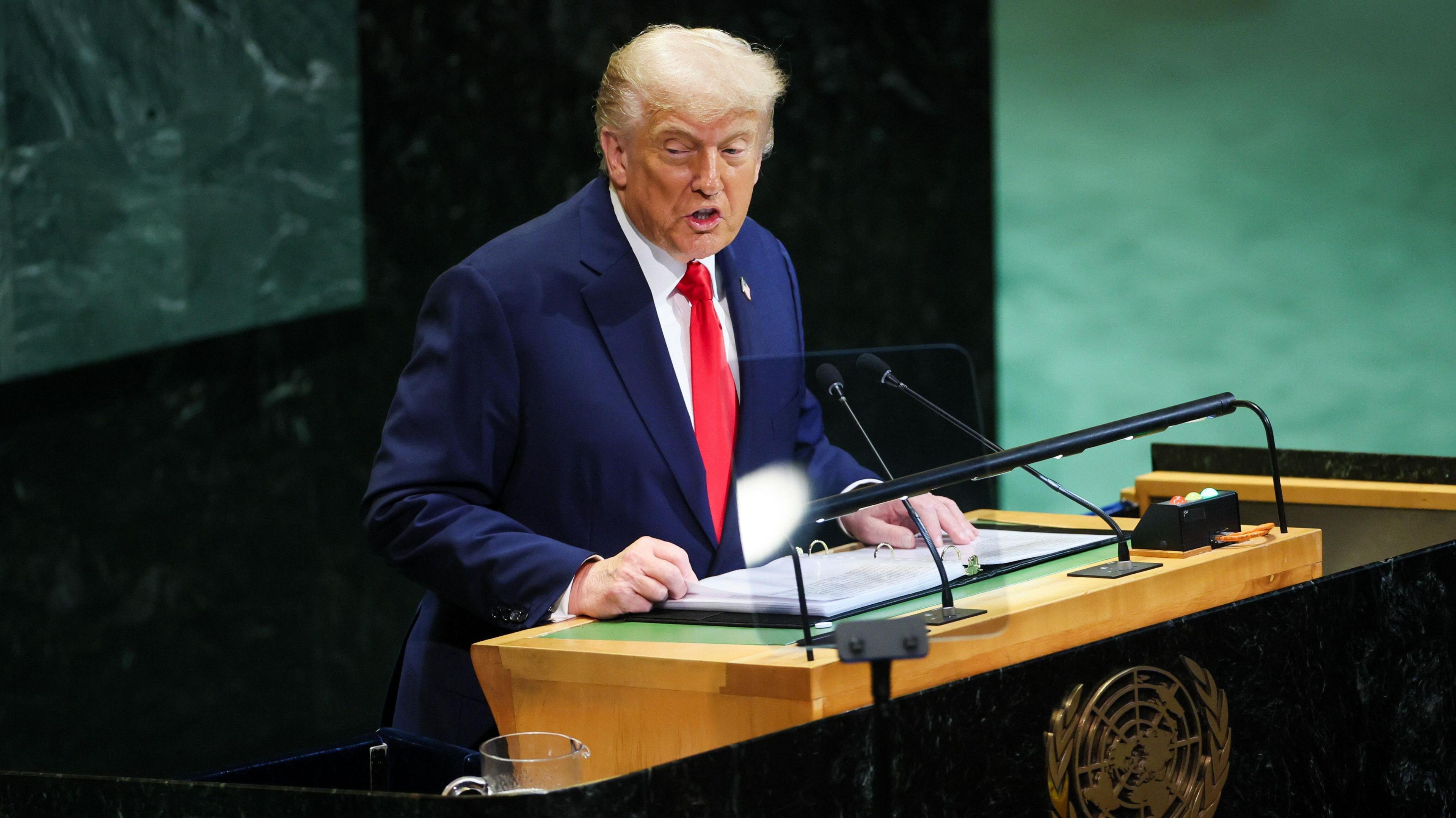 President Trump stands at the podium in the UN assembly hall in New York. The hall has a green carpet and green marble fittings. The president is wearing a blue suit and a red tie.