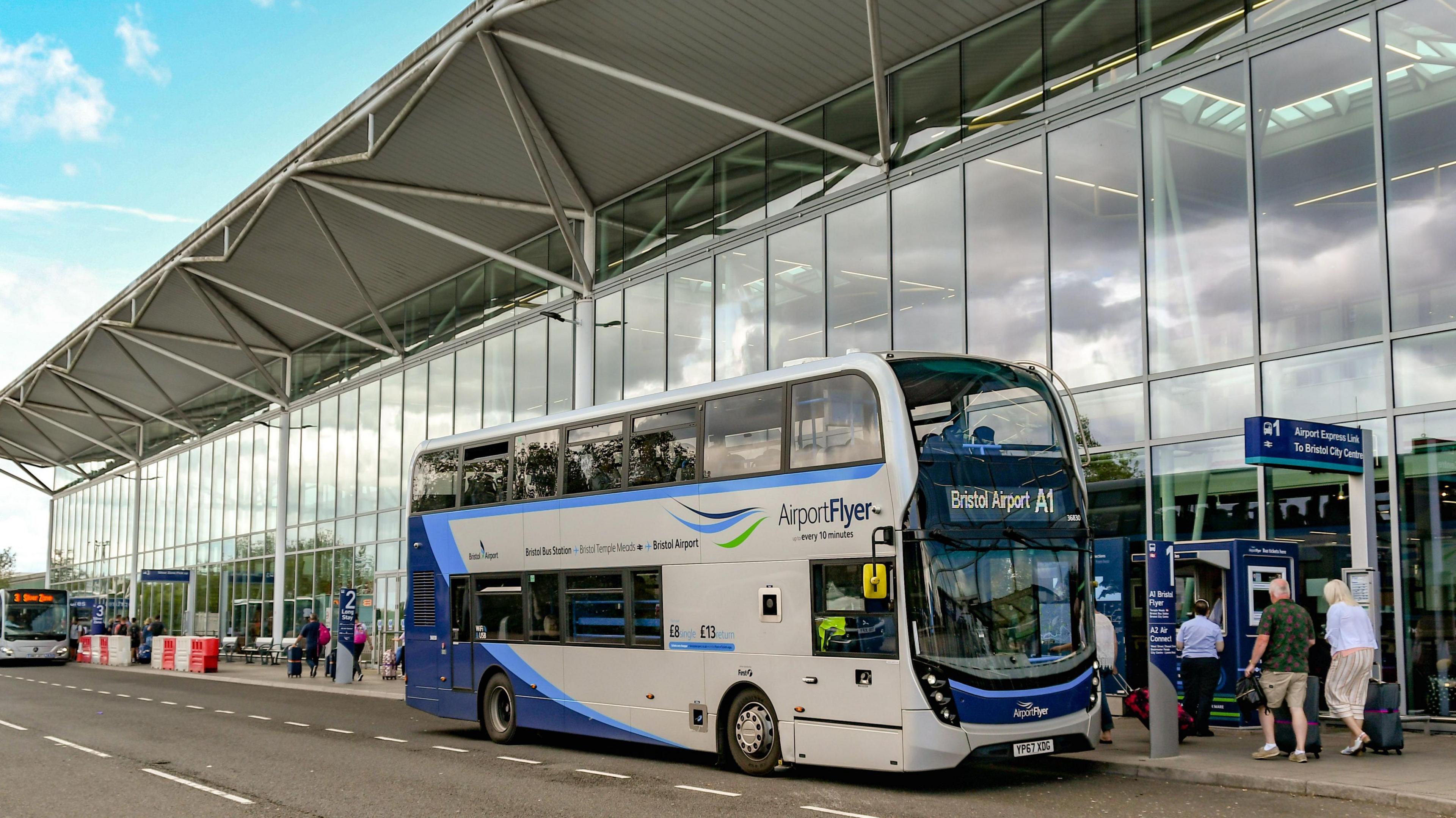 A view of Bristol Airport's terminal. The Airport Flyer bus is parked in front. Passengers can be seen with luggage.  