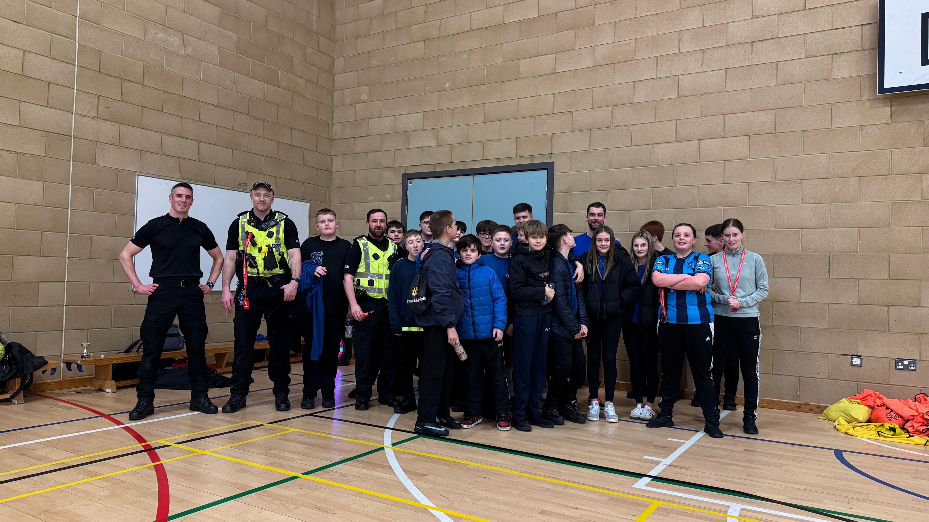 Cuid de na Poilis agus cuid de sgoilearan Sgoil MhicNeacail nan seasamh còmhla san talla spòrs agus iad air geama a chluich. 