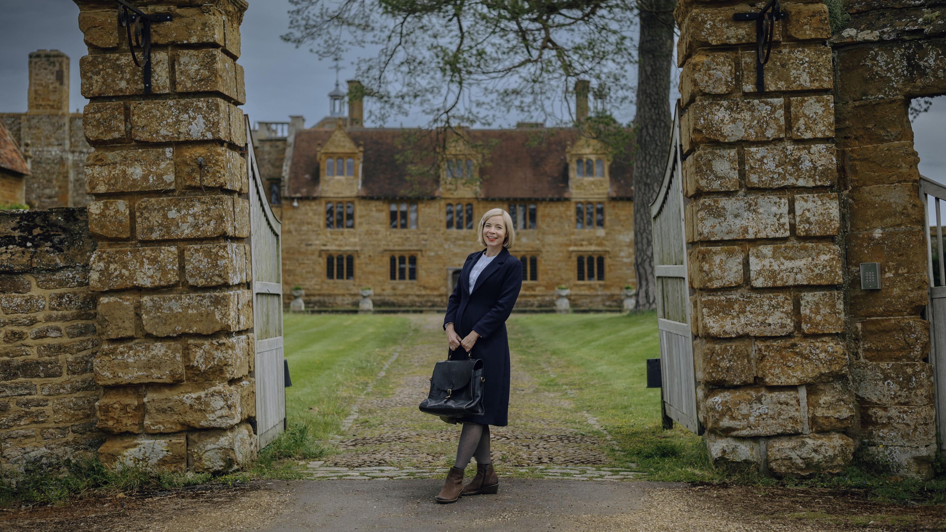 Lucy Worsley standing outside Ashby Manor, St Ledgers. She is holding a black bag, and wearing a dark coat, brown boots, is smiling and looking at the camera. She is standing between two wooden gates, brick columns, with a historical house behind her, with stone bricks and a large number of windows. 